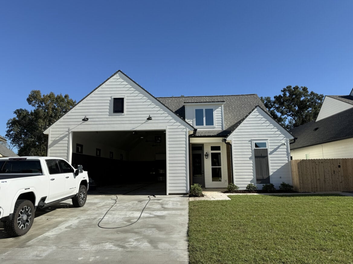 A white house with a double garage and gabled roof sits on a green lawn under a clear blue sky. A white pickup truck is parked in the driveway, and a garden hose lies on the concrete.