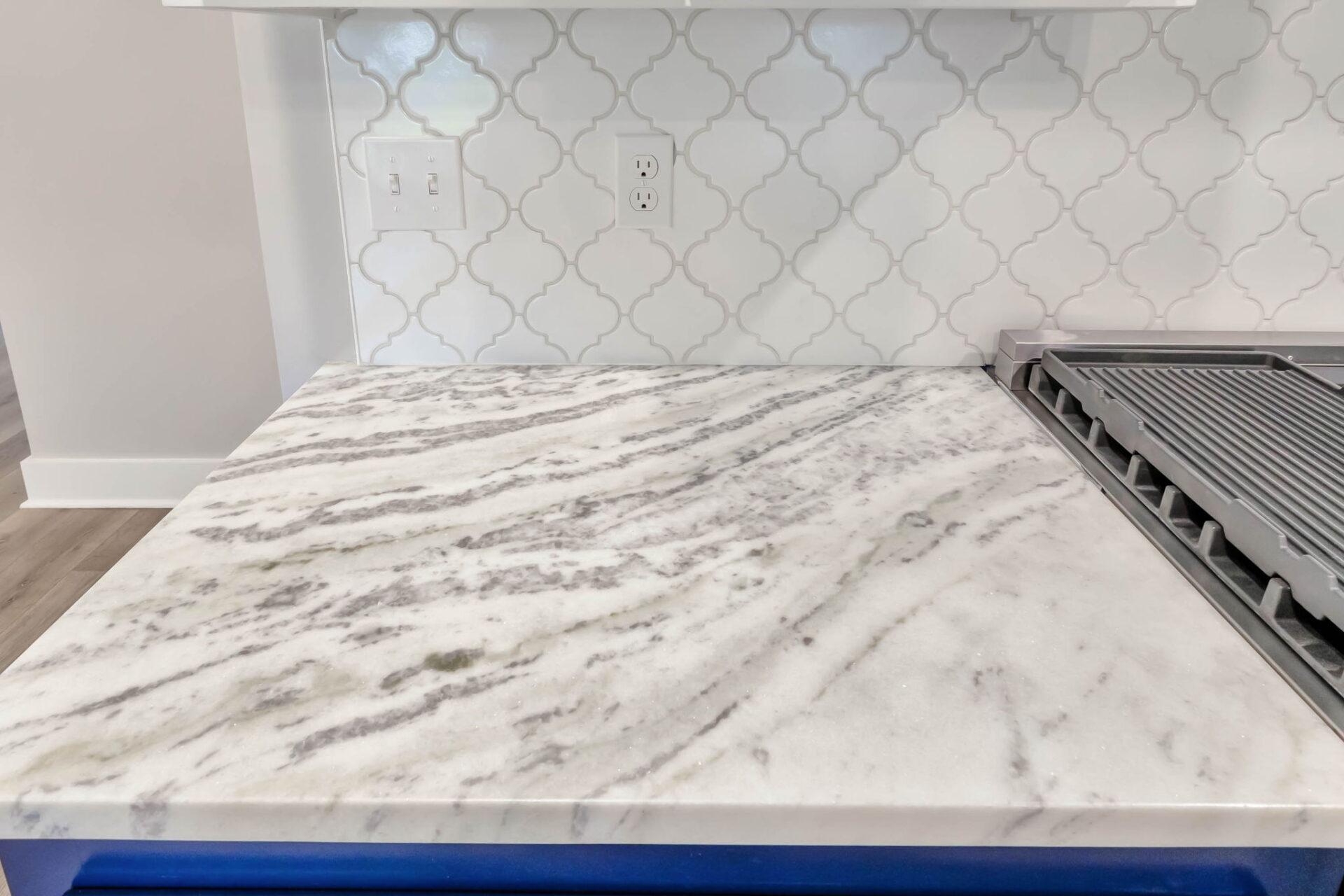 White and gray marble countertop next to a stovetop with a grill, set against a backsplash with a white geometric tile pattern. Two electrical outlets are visible on the backsplash above the counter.