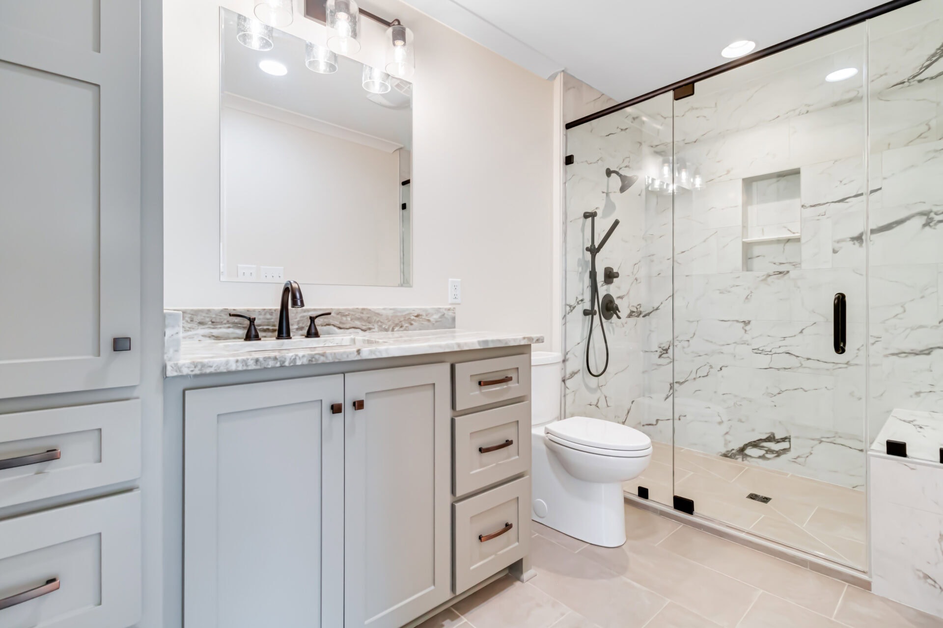 A modern bathroom with a double-light fixture above a rectangular mirror, light-colored vanity with drawers, a white toilet, and a glass-enclosed shower with marble walls and black fixtures.