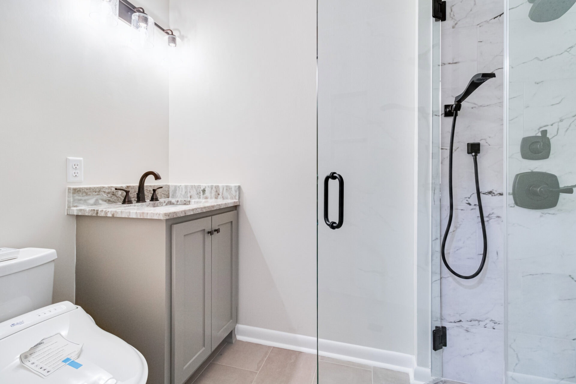 A modern bathroom with a white toilet, a gray vanity with a marble countertop and black faucet, light fixture above the mirror, and a glass-enclosed shower with a black showerhead and marble-style walls.