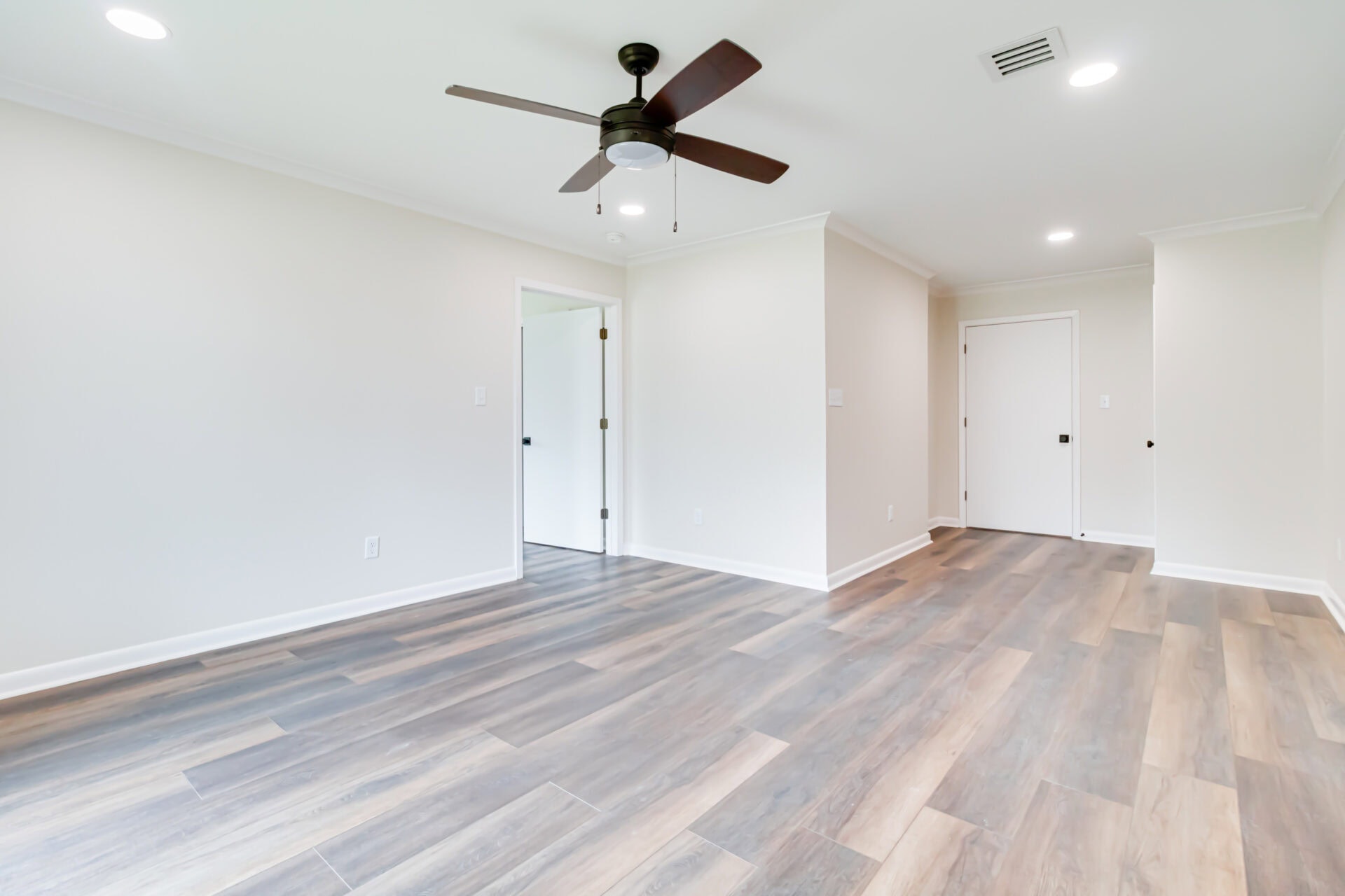 An empty room with light-colored walls, wood-look flooring, a ceiling fan with lights, white trim, and multiple doors. The room is well-lit with natural light from windows not visible in the image.