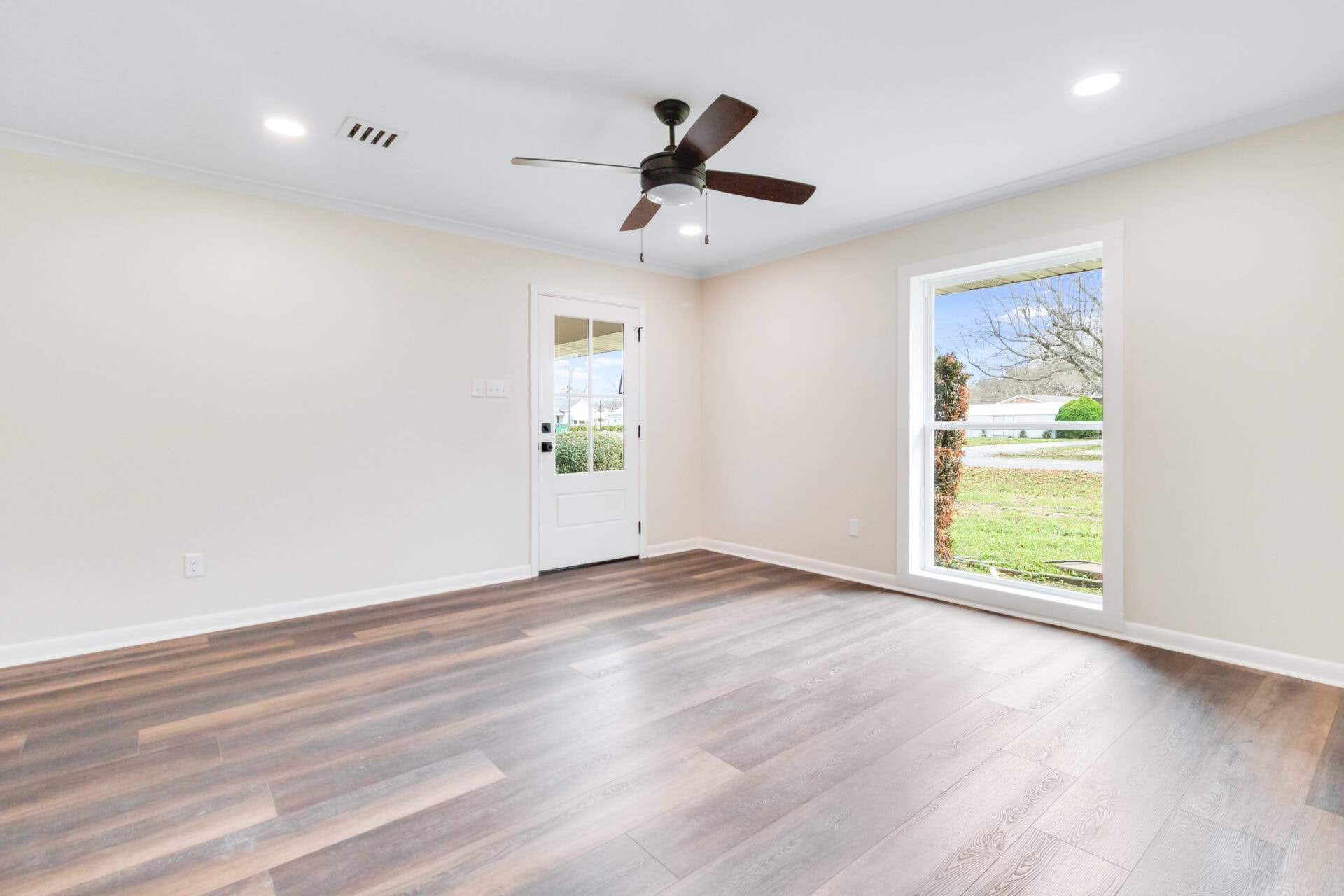 Empty room with light-colored walls, dark wood flooring, a ceiling fan with light, recessed lights, a white door with glass panes, and a large window showing grass and a tree outside.