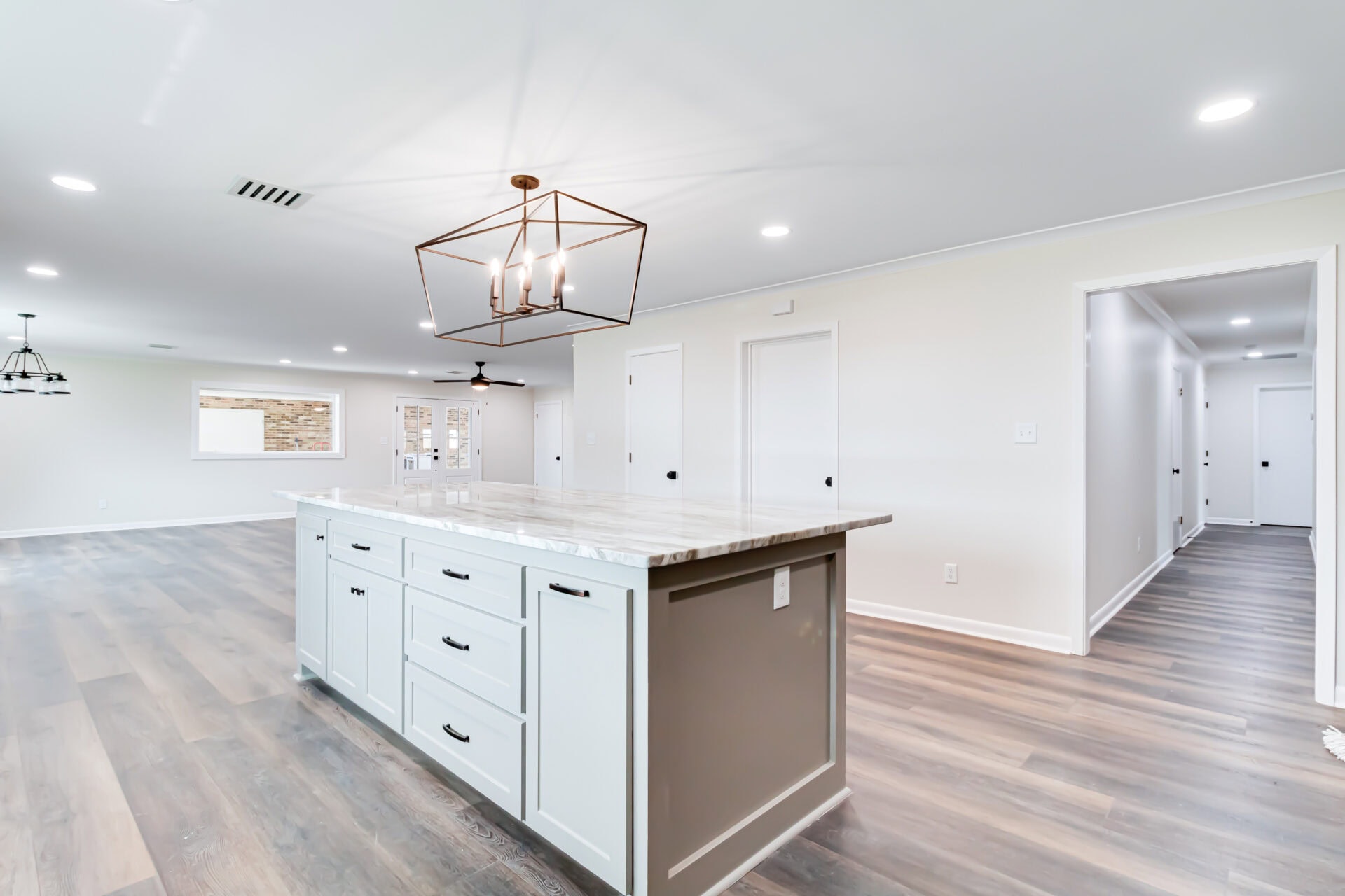 A modern kitchen with a large marble island, white cabinets, pendant lighting, and wood-look flooring. The space is open with multiple doors and a view of another room through a windowed wall.