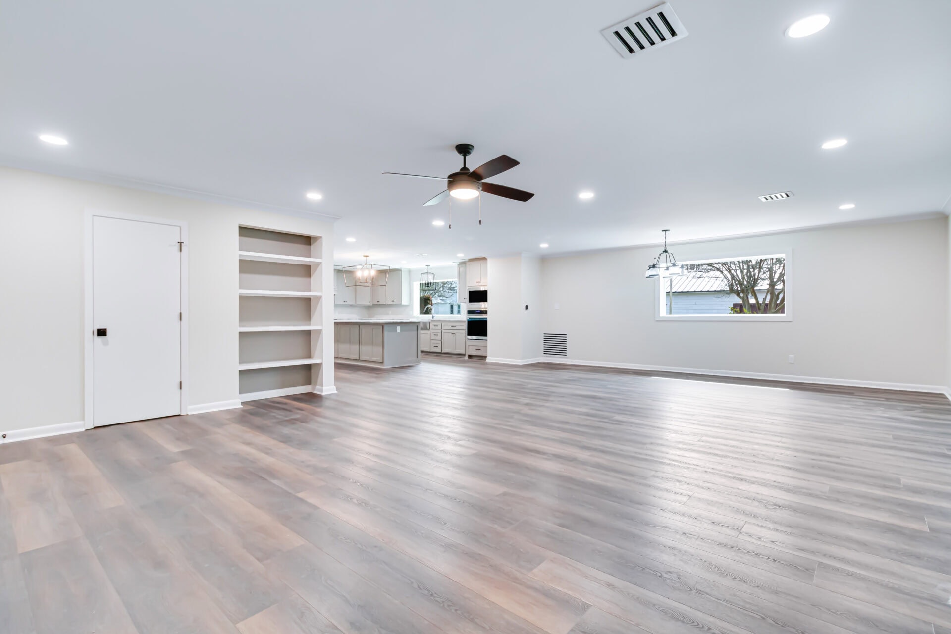 Spacious open-plan room with light wood flooring, recessed lighting, ceiling fan, built-in shelves, white walls, and a view into a modern kitchen with pendant lights and white cabinetry. Large window on one wall.