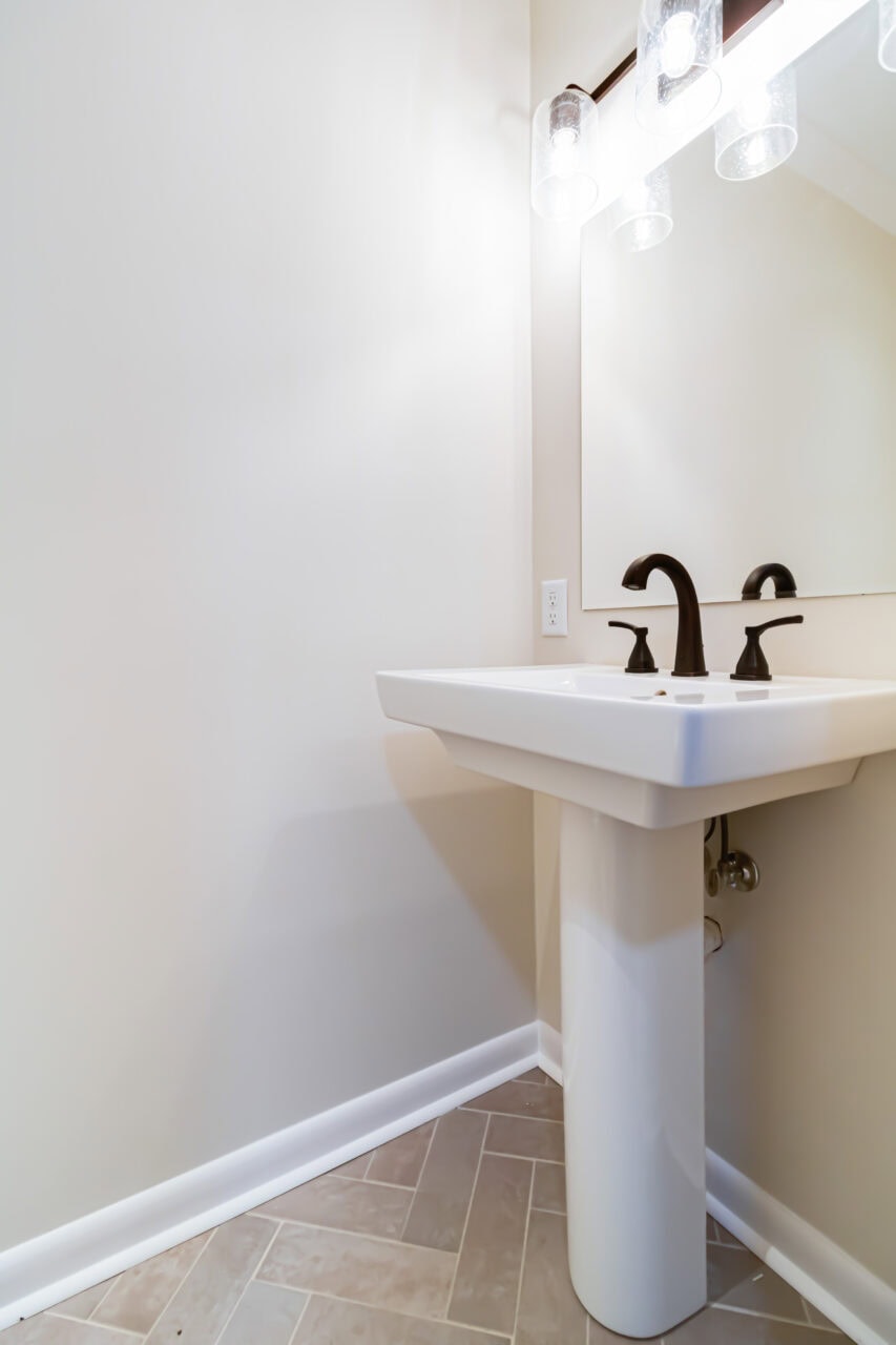 A white pedestal sink with black faucets is positioned in the corner of a bathroom. Above the sink is a rectangular mirror and three glass light fixtures. The walls are beige and the floor has a light herringbone pattern.