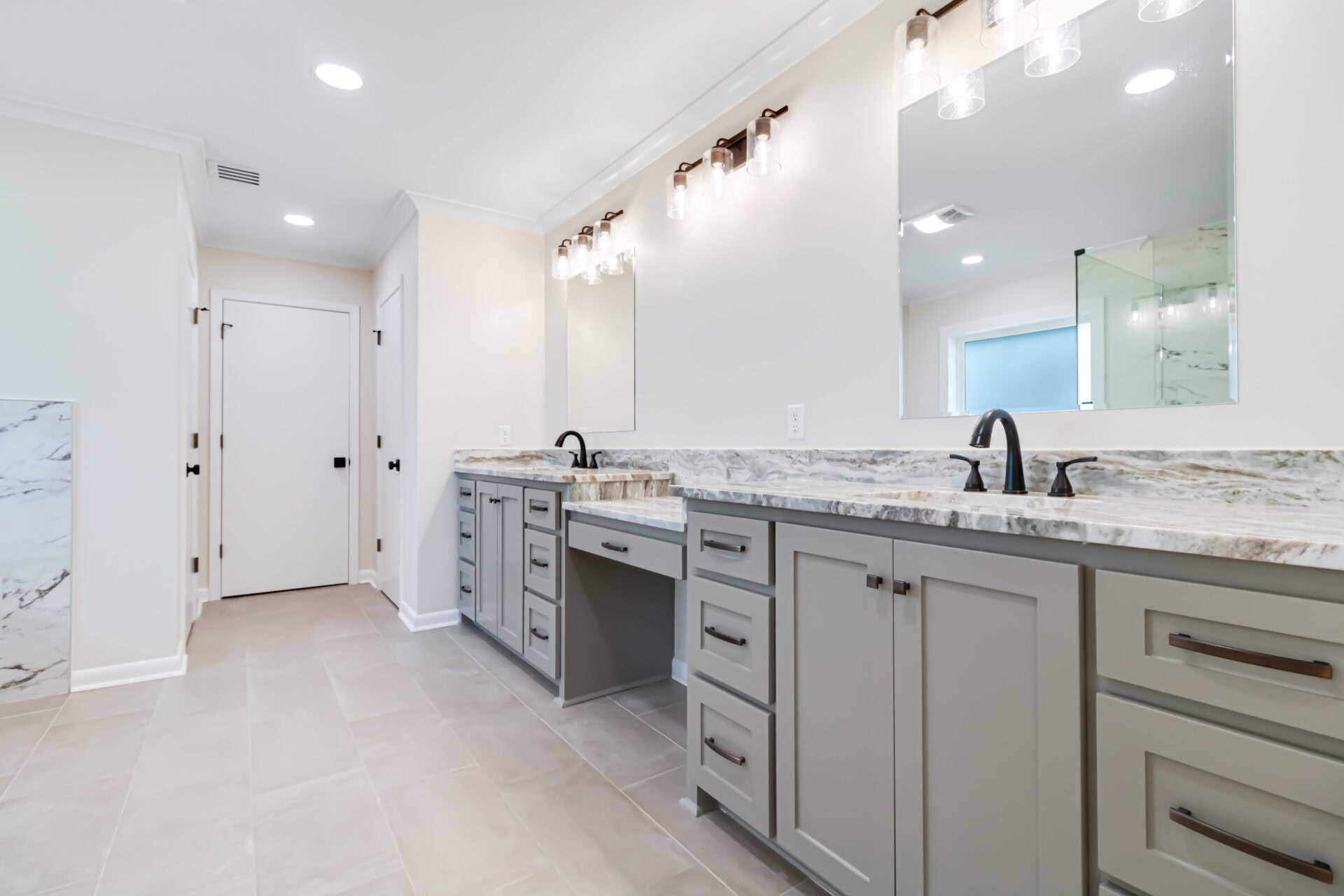 Modern bathroom with light gray cabinets, marble countertops, dual sinks with black faucets, large wall mirrors, and overhead lighting. Light tile flooring and neutral walls create a bright, spacious atmosphere.