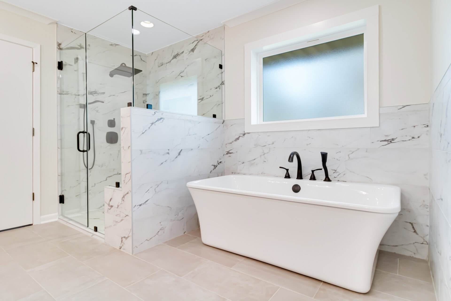 Modern bathroom with a glass-enclosed shower, white freestanding bathtub, marble walls, matte black fixtures, a frosted window, and beige tile flooring.