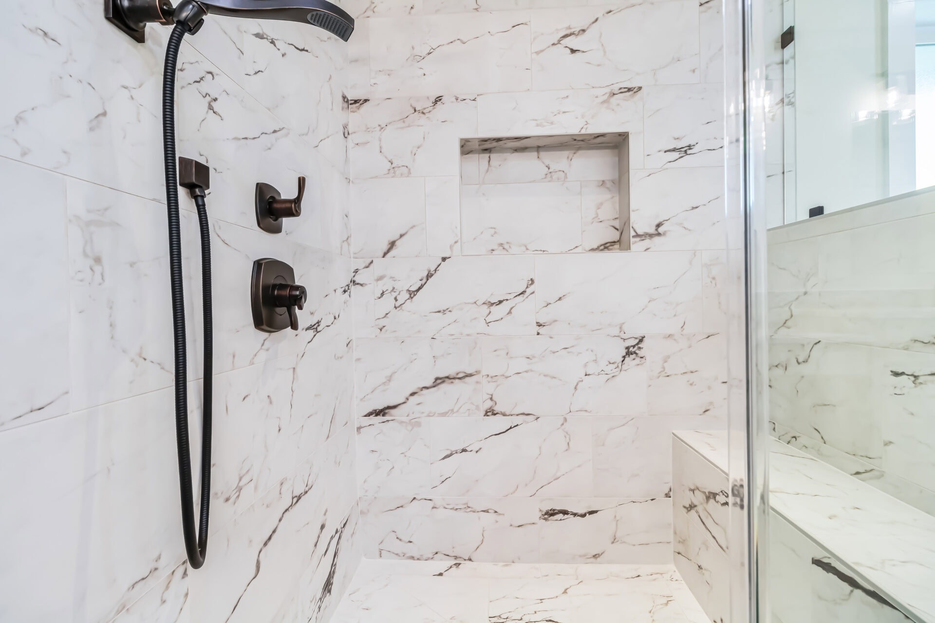 Shower with white and gray marble tile walls, built-in shelf, black showerhead and handles, and a bench on the right side. A glass shower door is partially visible.