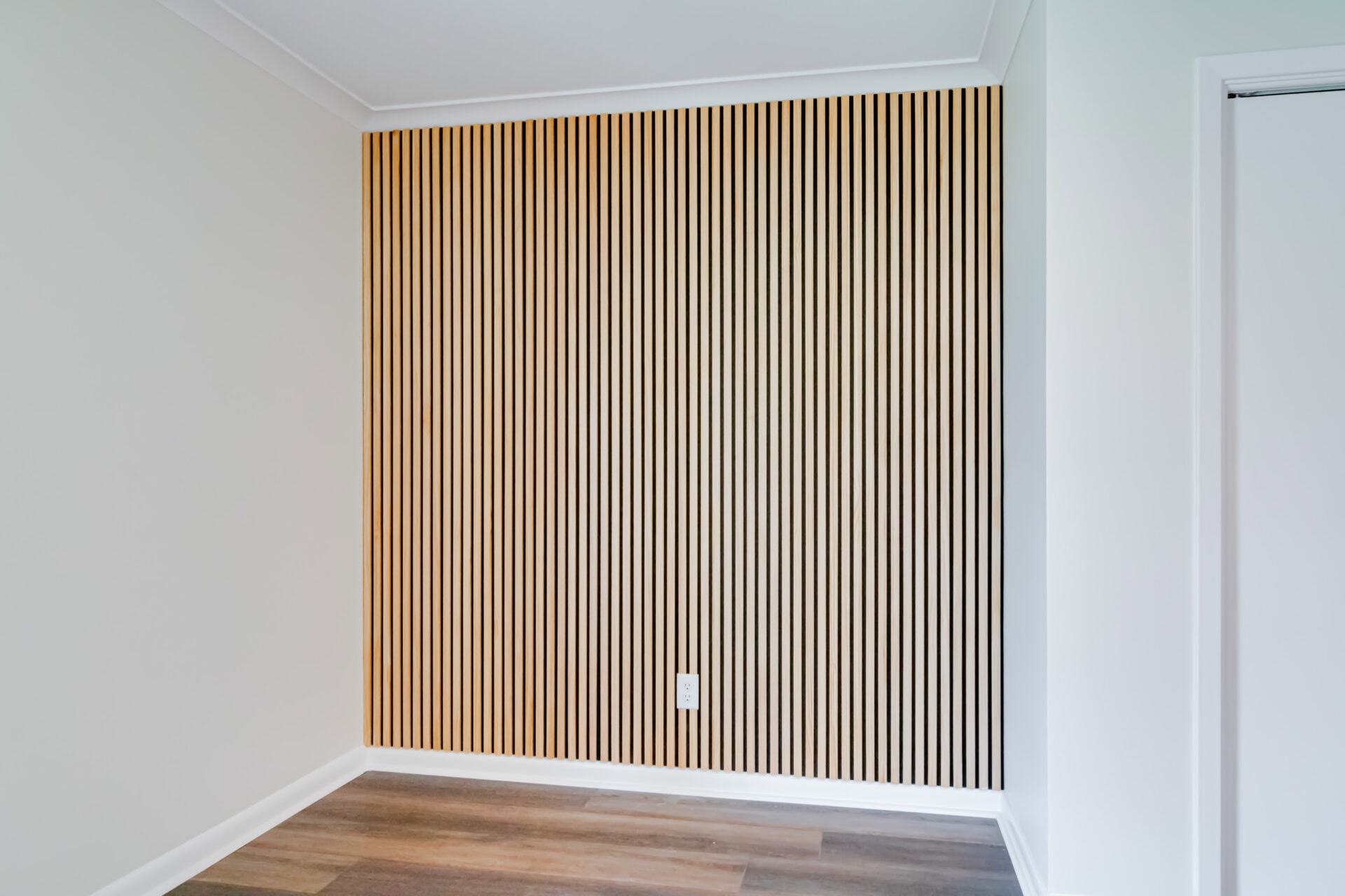 A modern interior wall with vertical wooden slats, a white electrical outlet at the bottom center, white trim, and adjacent white walls. The floor is covered with wide, dark wood planks.