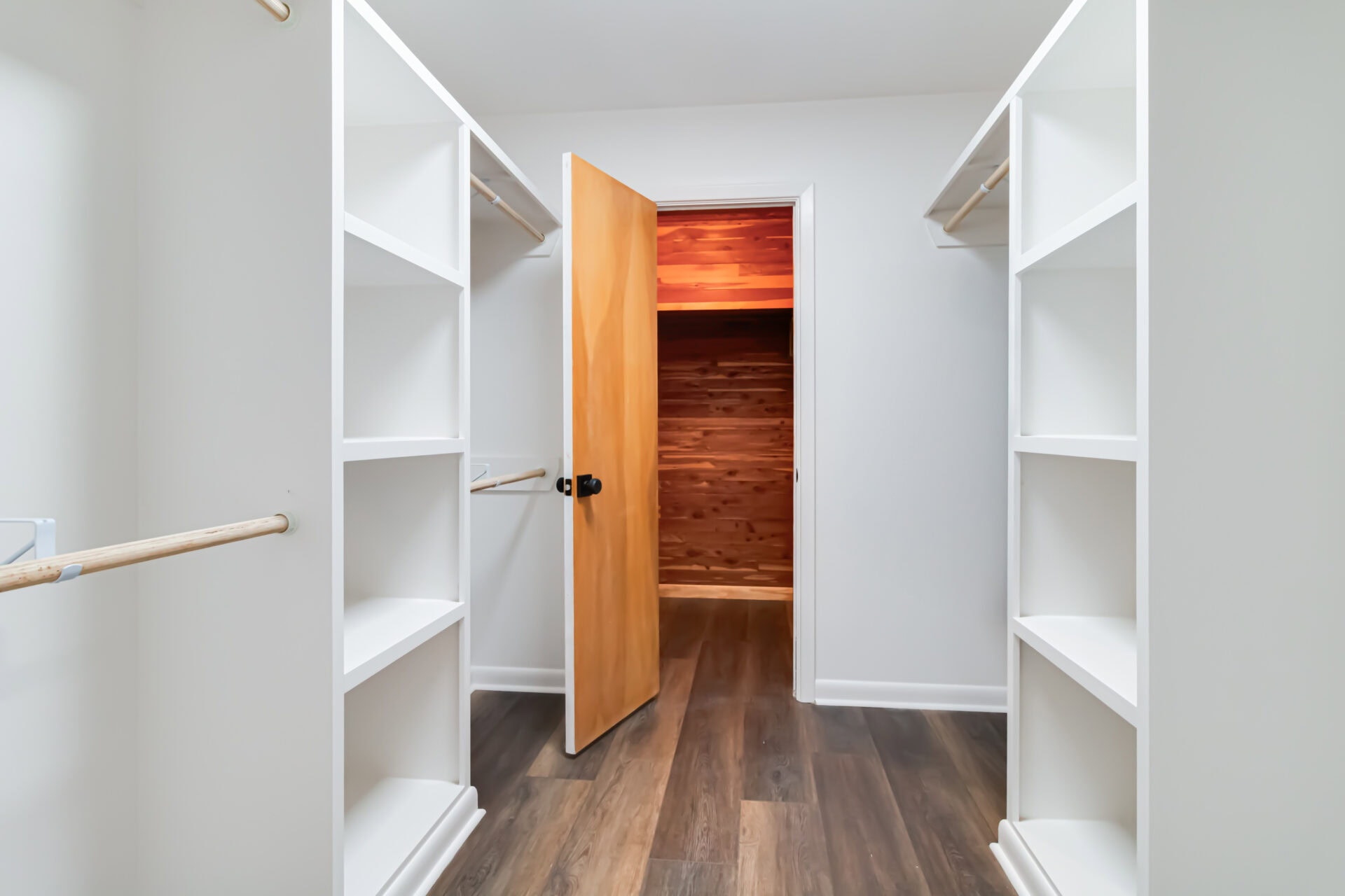 A walk-in closet with white built-in shelving and hanging rods on both sides, dark hardwood floors, and an open wood door leading to a cedar-lined storage room.