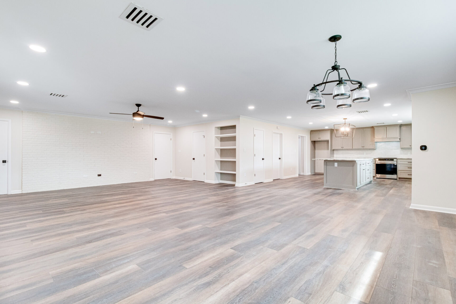 Spacious open-concept living area with light wood flooring, built-in shelves, ceiling fans, recessed lighting, and a modern kitchen with an island and pendant lighting in the background.