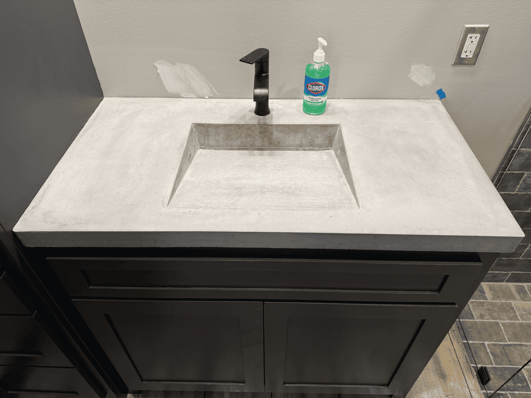 A gray concrete bathroom sink with a built-in rectangular basin, a black faucet, and a bottle of Clorox soap on the countertop. There are some wall patches above the sink and a visible electrical outlet.
