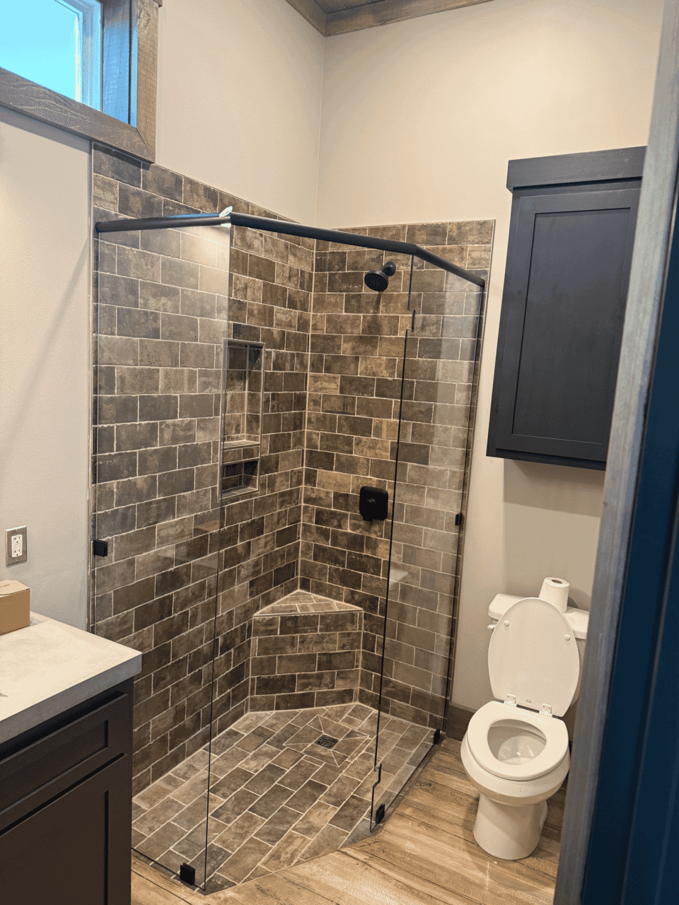 A bathroom with a glass-enclosed, corner shower with brown tile walls and floor, a white toilet with the lid up, a dark vanity, a wall cabinet, and a small high window.