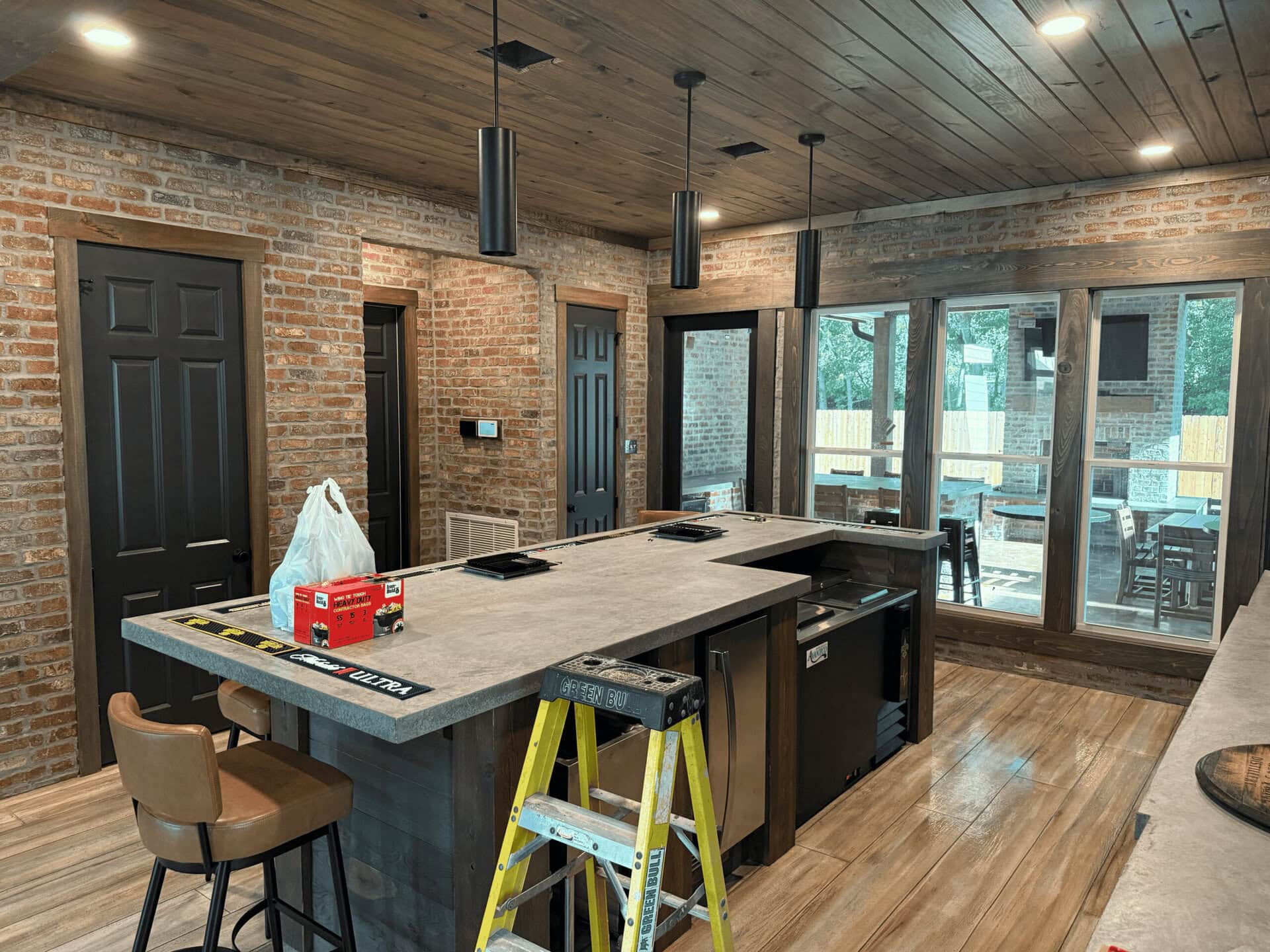 A kitchen with exposed brick walls, wooden ceiling and floor, a large island with a sink, and black cabinets. A step ladder, bar stools, a ruler, and a plastic bag are on or near the island. Large windows are in the background.