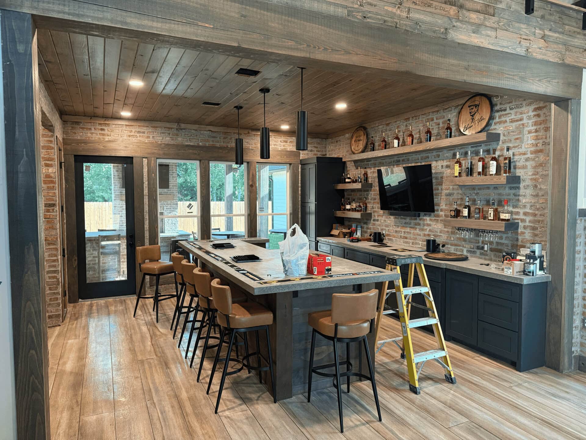 A modern home bar with brick walls, wooden ceiling and floor, shelves with bottles, a mounted TV, a counter with barstools, a stepladder, and groceries and boxes on the counter. Large windows and a door lead outside.