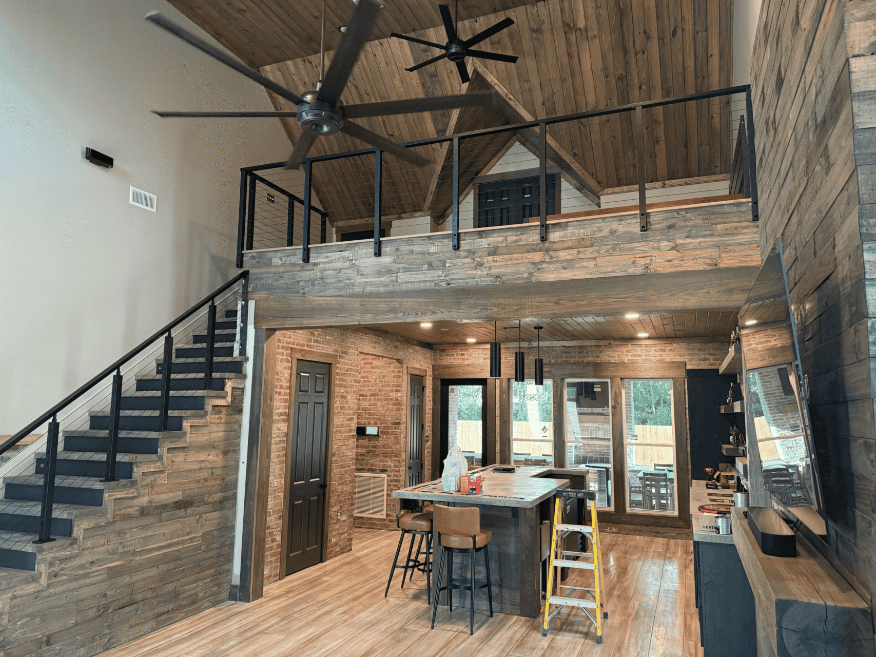 A modern rustic kitchen and living area with wood-paneled walls, an island, black metal railings on a staircase, a loft, ceiling fans, a ladder by the island, and large windows letting in natural light.
