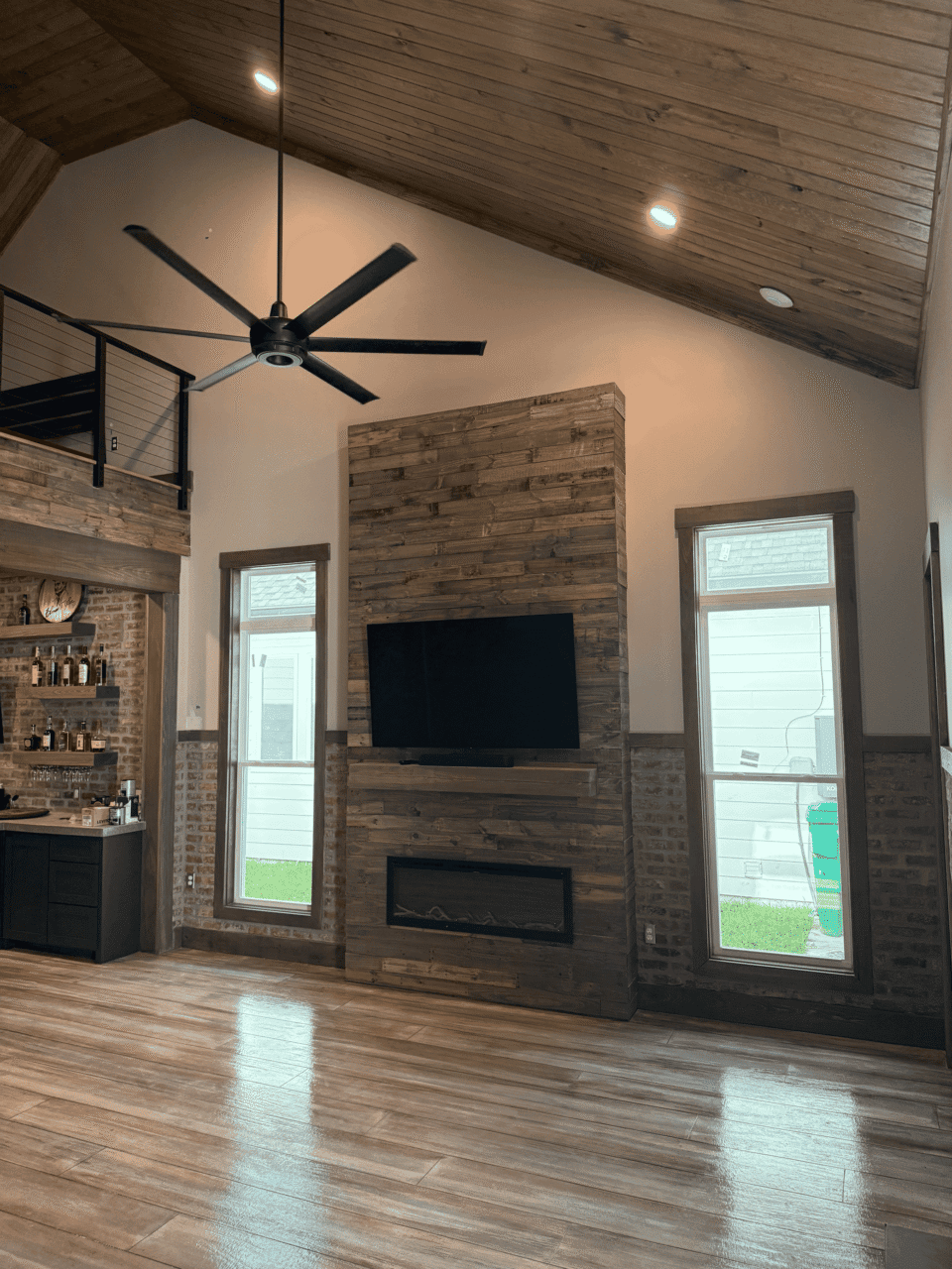 A living room with wood-paneled walls and ceiling, featuring a mounted TV above a modern fireplace between two tall windows. A black ceiling fan hangs from the vaulted ceiling.