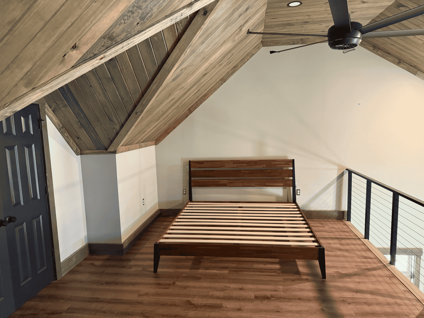 A loft bedroom with wood-paneled sloped ceiling, an empty wooden bed frame, a ceiling fan, wood floor, and a dark door; the area is lit with natural light and bordered by a metal railing.