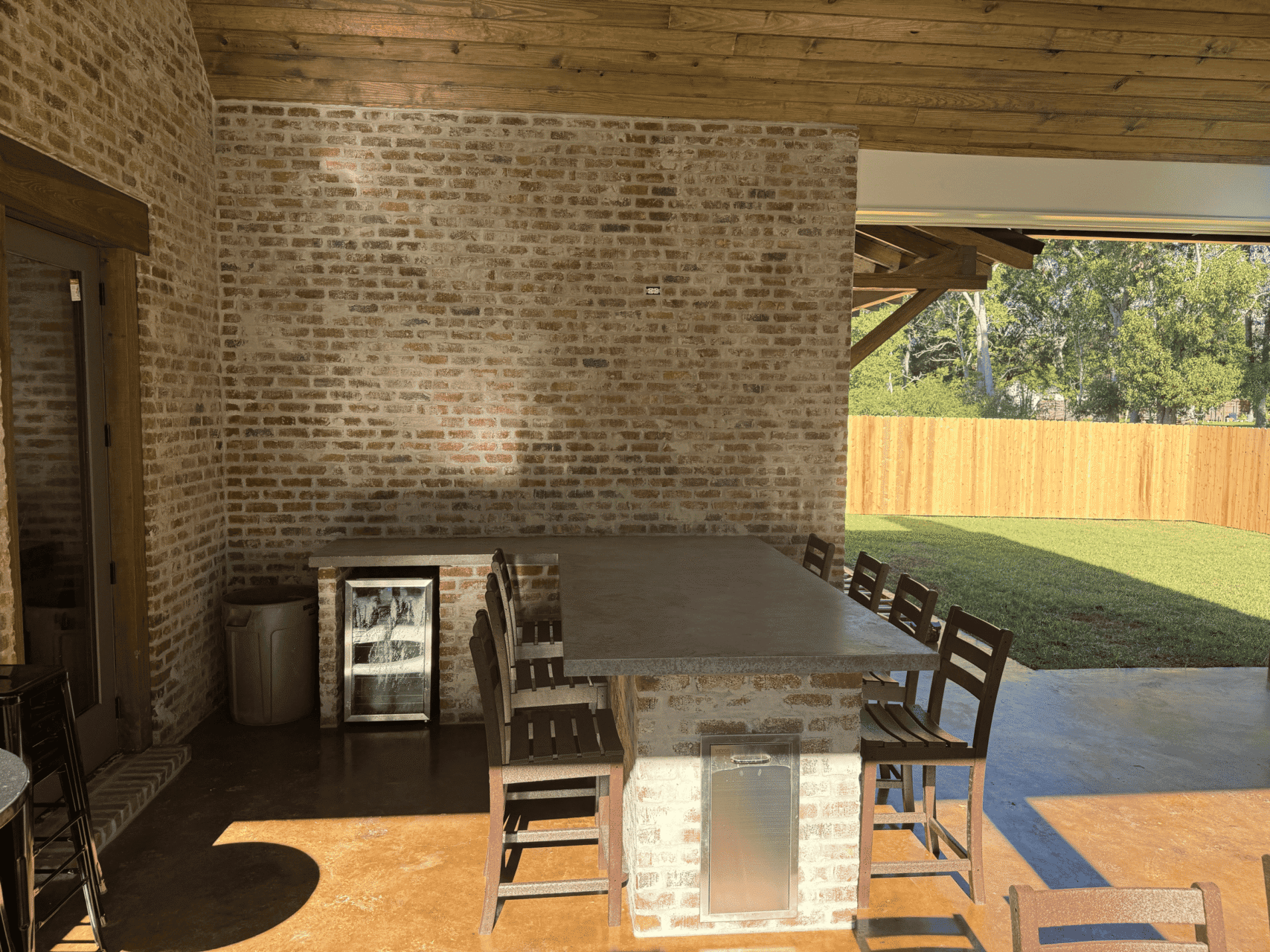 An outdoor patio with a brick bar area, several wooden bar stools, a mini refrigerator, and a trash can, under a wooden roof, next to a grassy yard with a wooden fence.