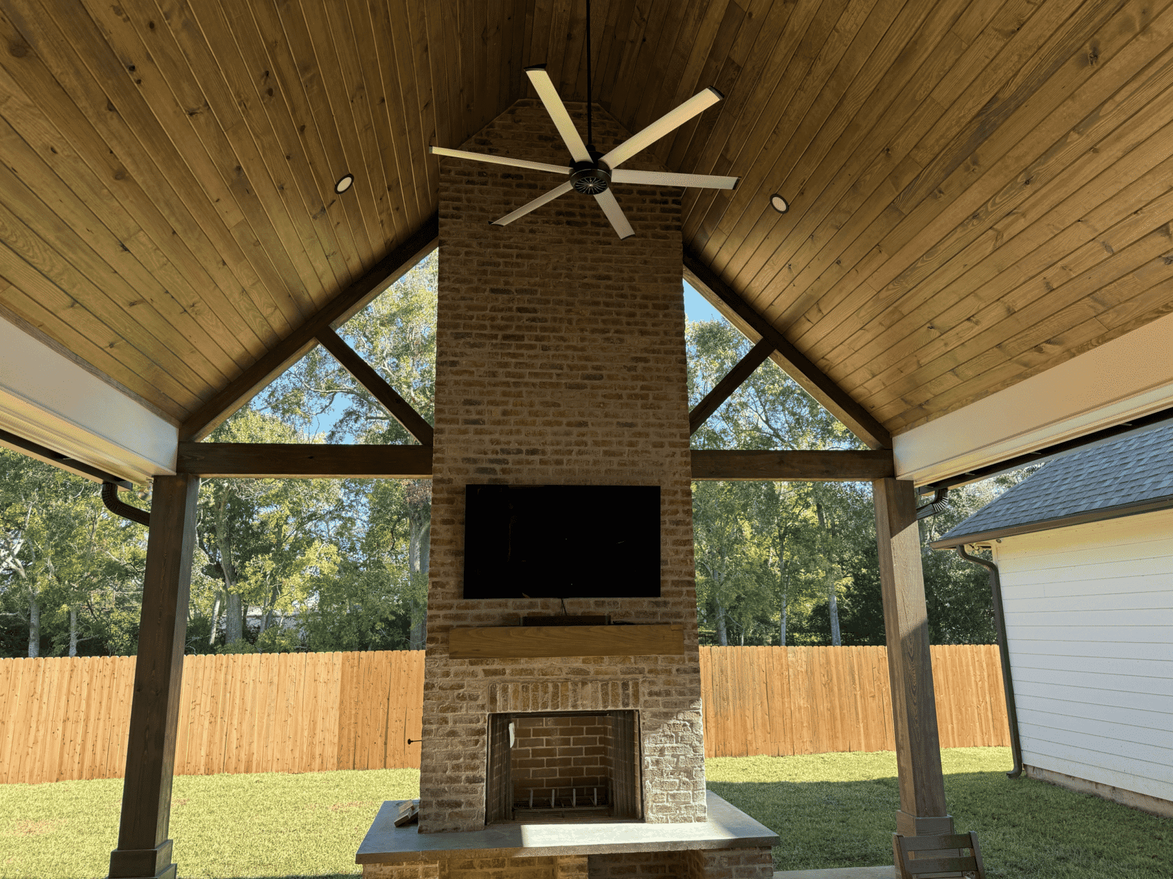 A covered outdoor patio with a vaulted wooden ceiling, a large brick fireplace with a TV mounted above it, and a ceiling fan. The space overlooks a fenced backyard with trees in the background.