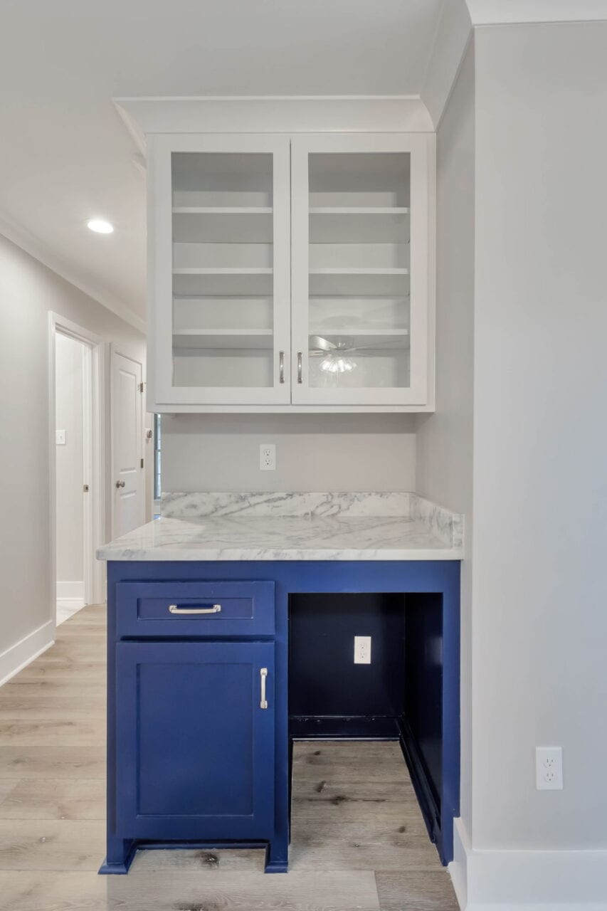 A small kitchenette area with a blue lower cabinet, a white marble countertop, and white upper cabinets with glass doors. The wall and trim are light gray, and the floor is light wood. An open space under the counter reveals outlets.