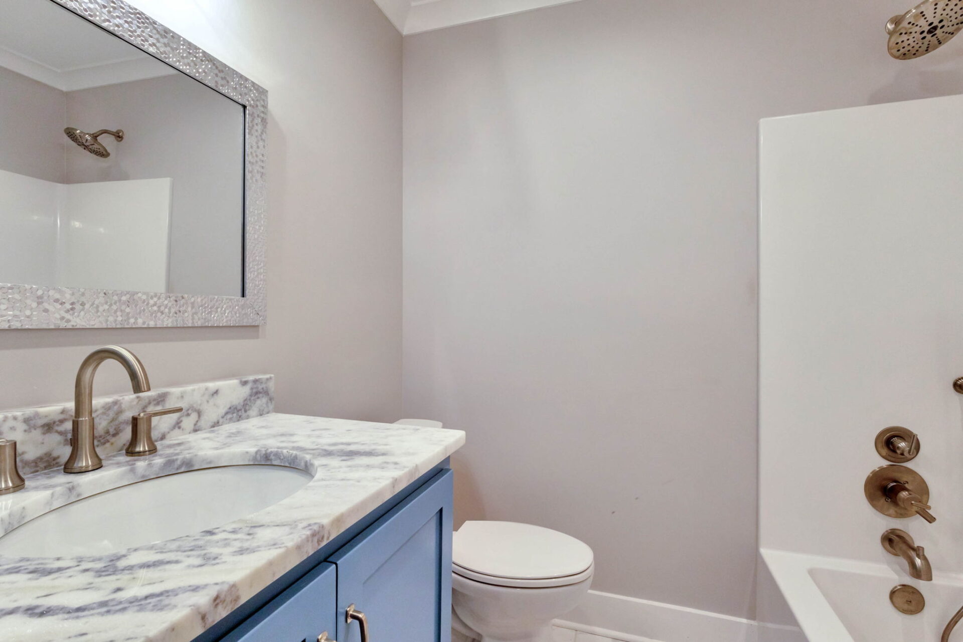 A bathroom with a marble countertop, blue vanity, large wall mirror, silver faucet, toilet, and a bathtub with a wall-mounted showerhead and fixtures. The walls are light gray.