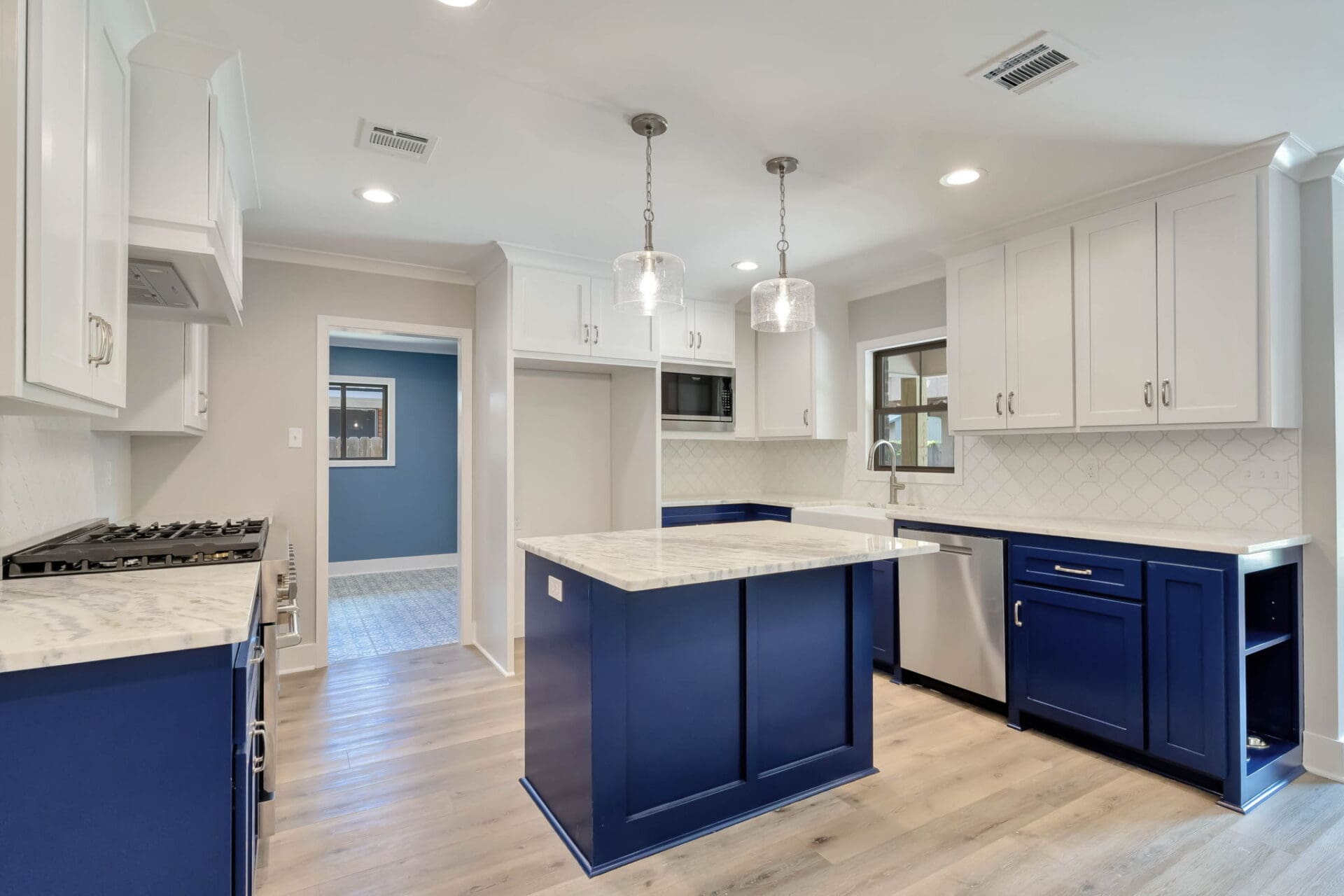 Modern kitchen with white upper cabinets, blue lower cabinets and island, marble countertops, stainless steel appliances, light wood flooring, and two pendant lights over the island.