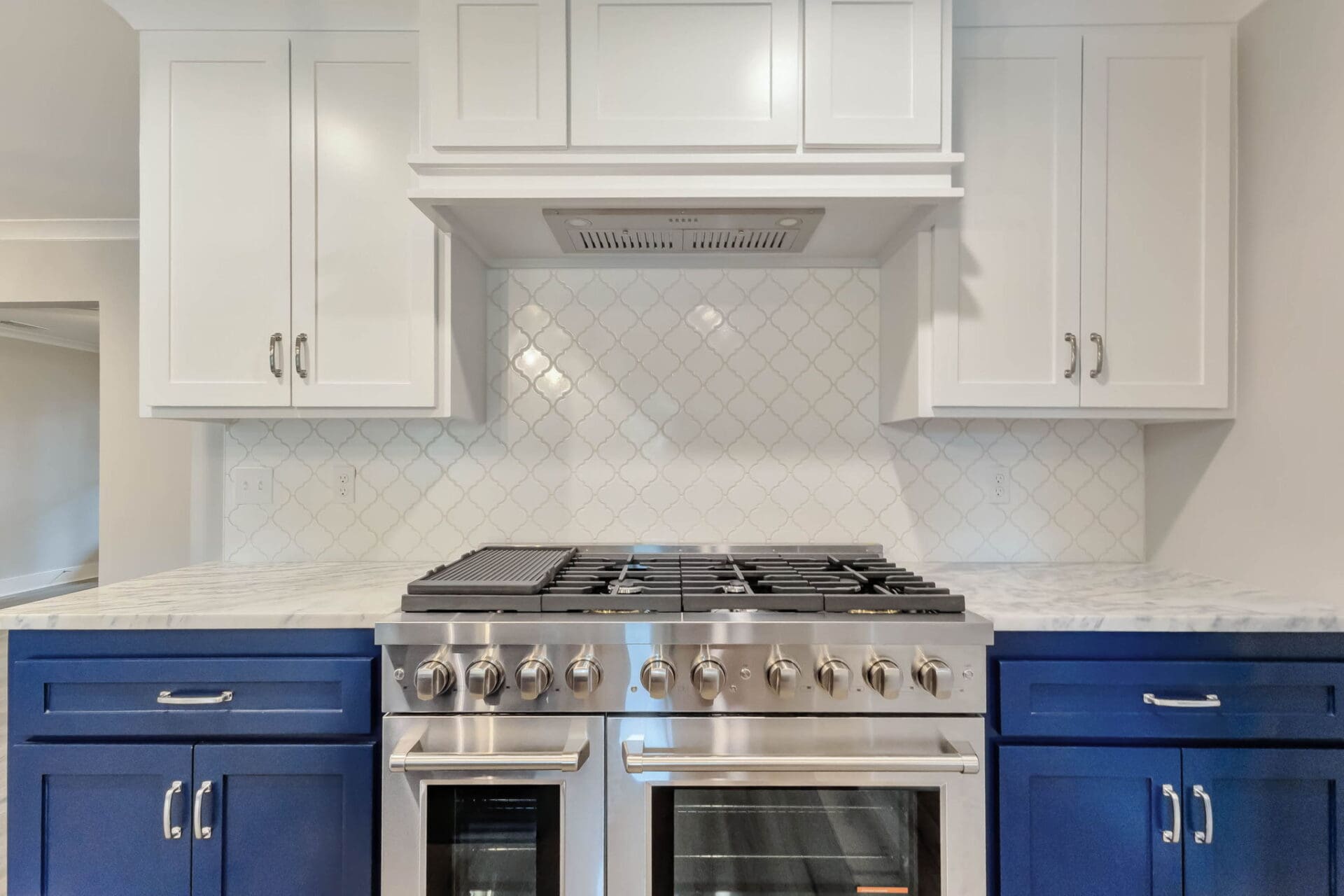 A stainless steel gas range with two ovens is set between blue lower cabinets and topped with white countertops. White upper cabinets and a white tile backsplash are also visible.
