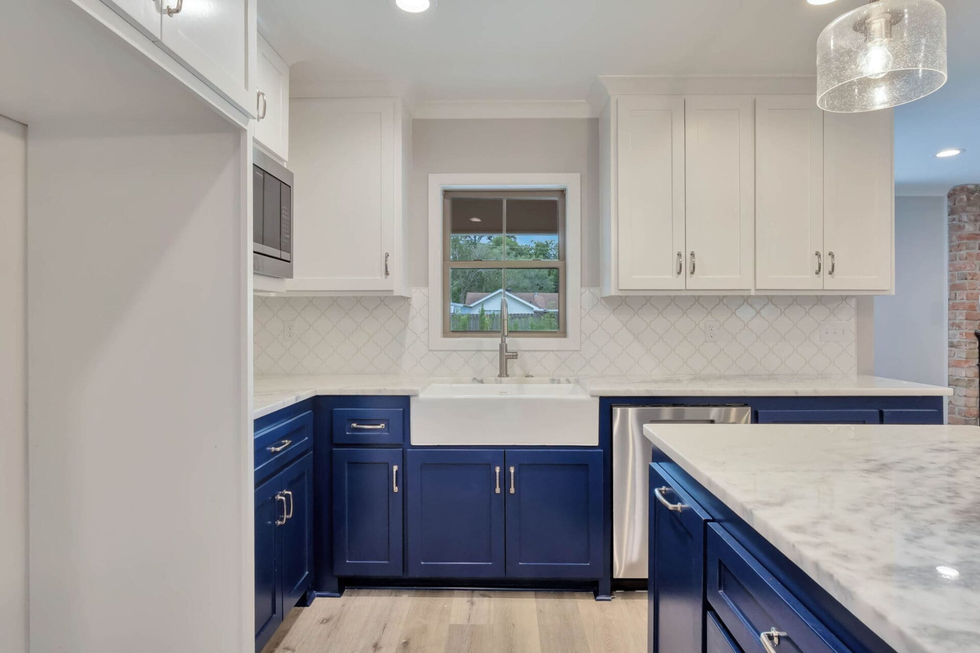 A modern kitchen with blue lower cabinets, white upper cabinets, a farmhouse sink under a window, marble countertops, a white tile backsplash, and wood flooring.