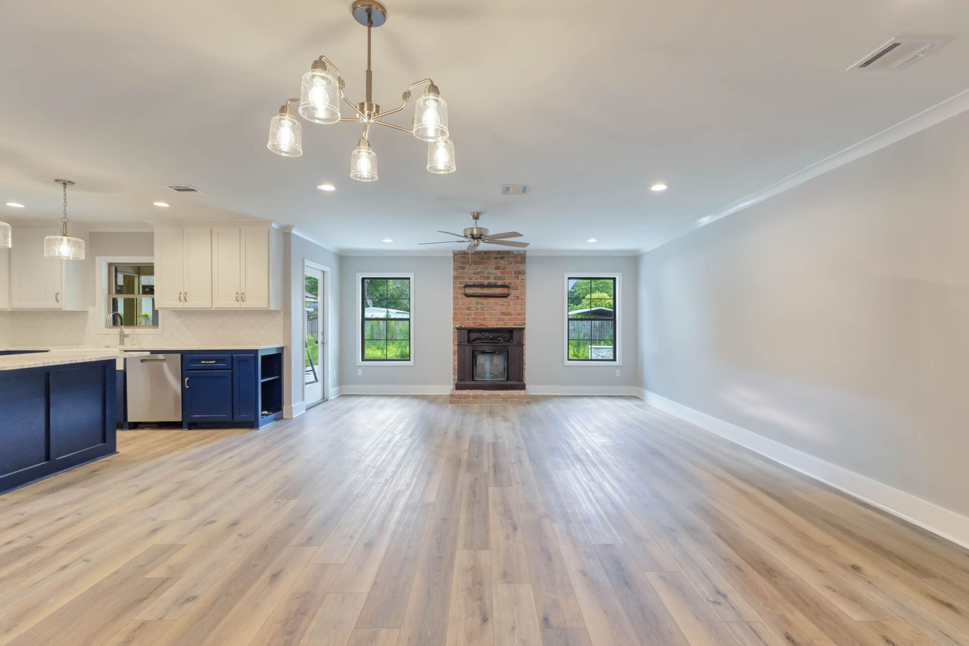 Spacious open-concept living area with light wood flooring, a brick fireplace, large windows, and a modern kitchen with white and blue cabinets, pendant lights, and marble countertops.