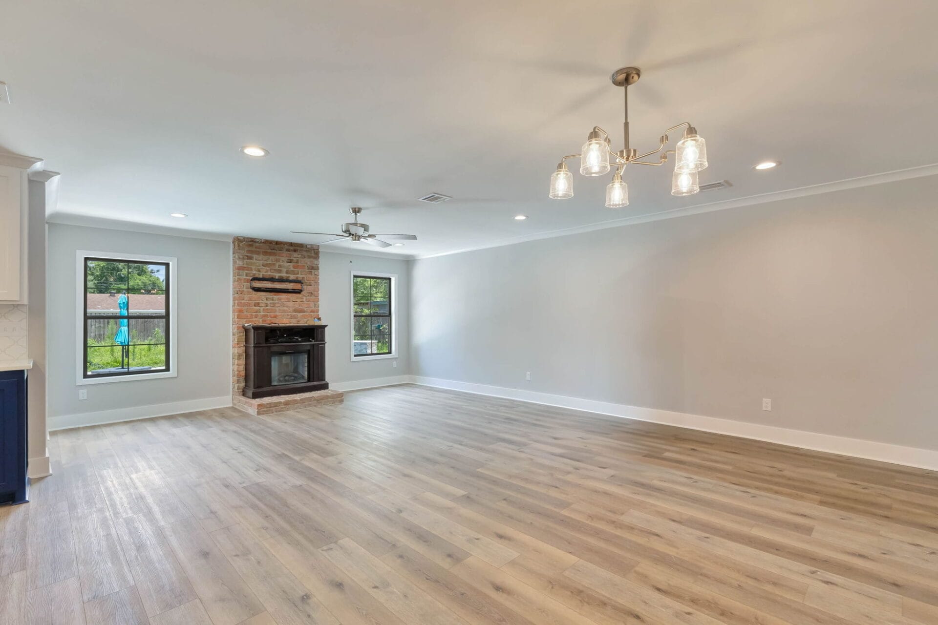 Spacious, empty living room with light wood flooring, a brick fireplace, ceiling fan, modern chandelier, and two large windows overlooking a backyard. Walls are light gray with white trim.