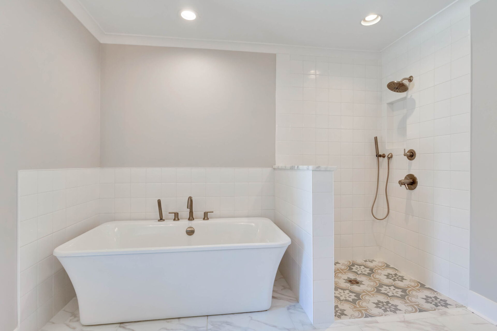 A bathroom with a white freestanding tub on the left and an open walk-in shower on the right featuring white tiles, patterned floor tiles, and gold fixtures. The space has recessed ceiling lights and neutral walls.