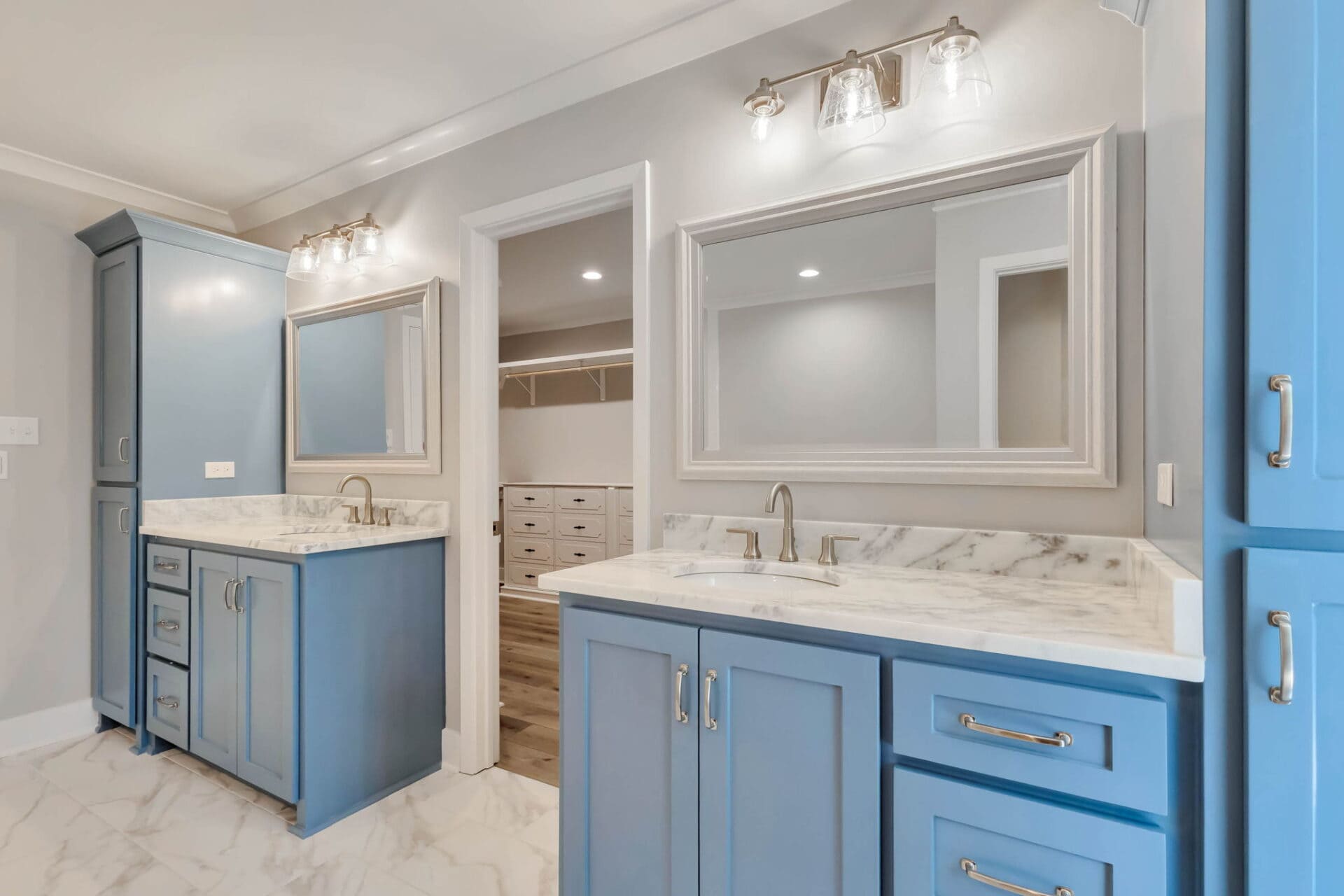 A bathroom with two blue vanities, each with a sink, marble countertop, and a large mirror above. There are two light fixtures above each mirror and white marble floor tiles. A walk-in closet is visible through an open doorway.