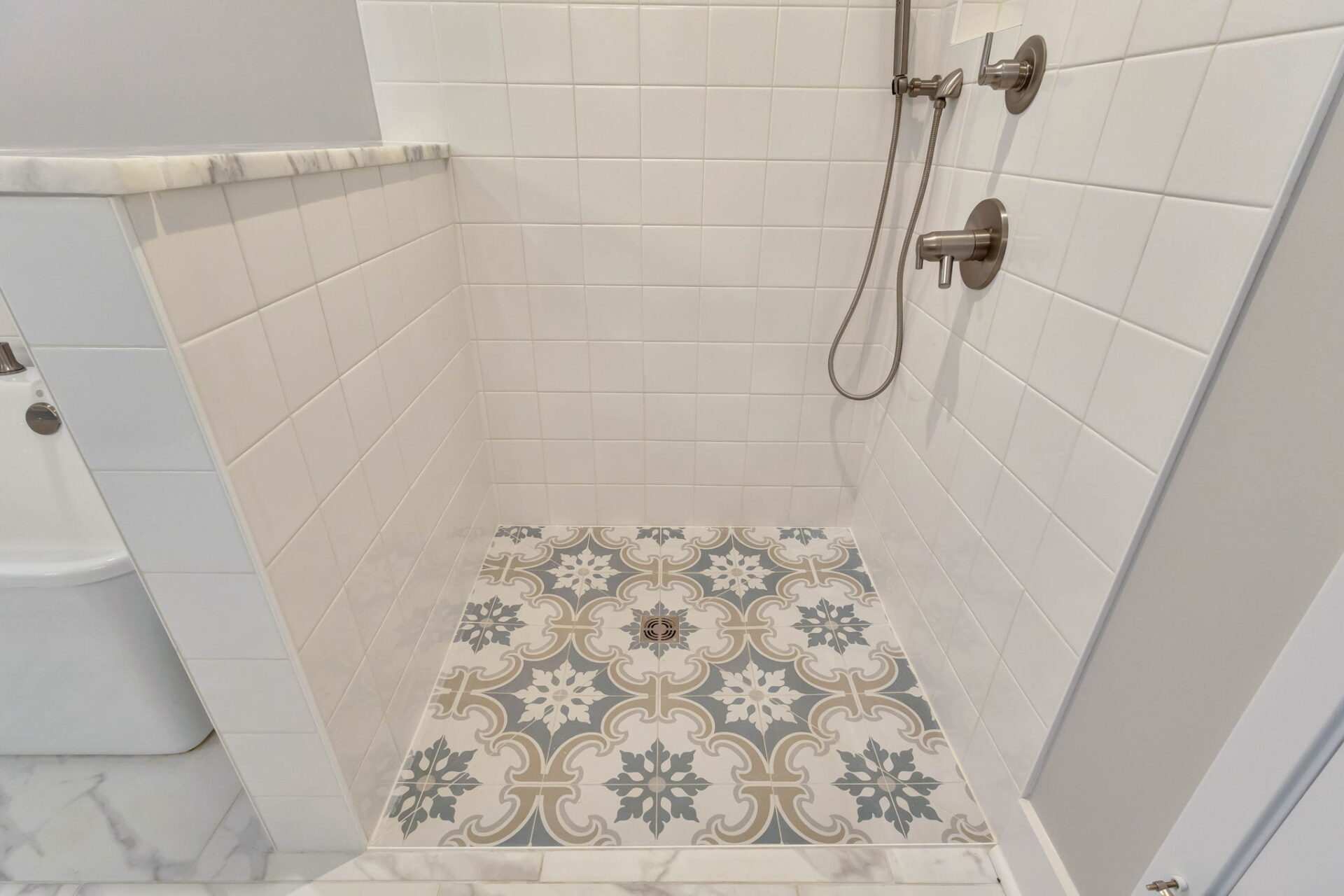 A shower with white square tiles on the walls, a handheld showerhead and control knobs, and a floor with patterned tiles in gray, beige, and white geometric designs.
