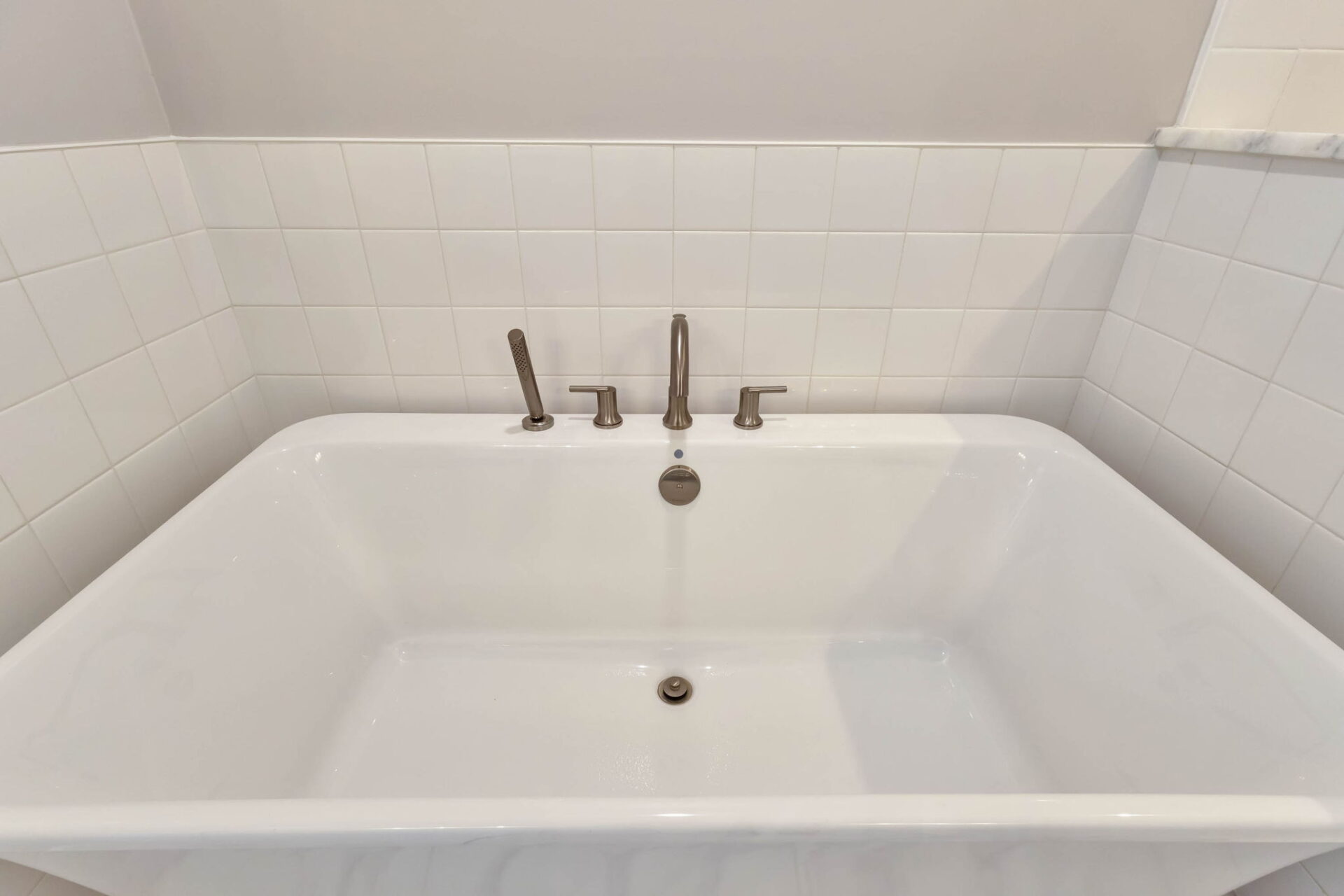 A white rectangular bathtub with metal faucet and handles is shown installed against a tiled wall in a bathroom. The tub is clean and empty, with a central drain and white tile backsplash.