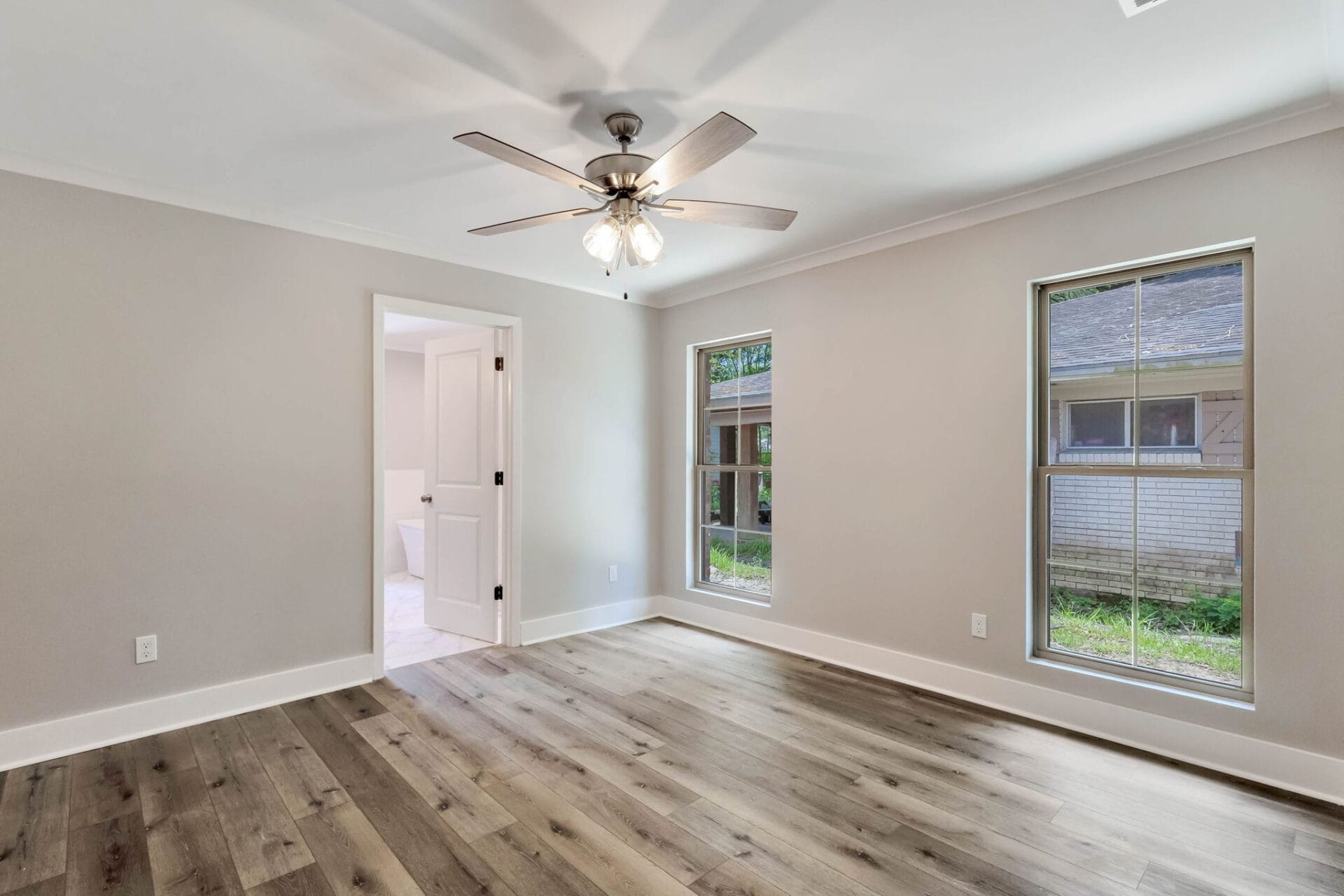 Empty room with light gray walls, wood flooring, ceiling fan with lights, two tall windows, and an open door leading to a bathroom; view outside shows a building and greenery.