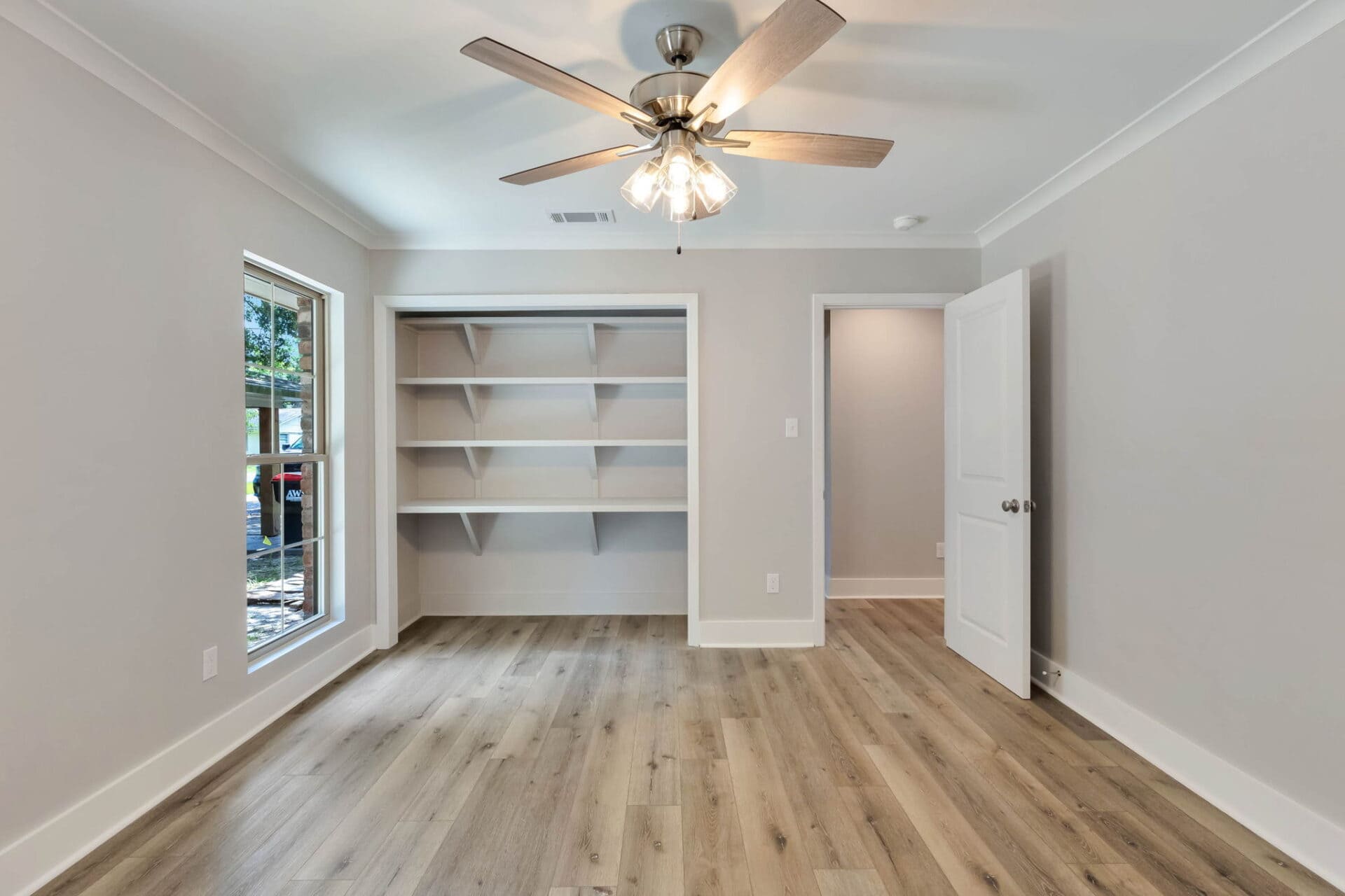 A bright, empty room with light wood flooring, a ceiling fan with lights, built-in shelves in an alcove, a large window on the left, and an open door on the right. Walls are painted light gray.