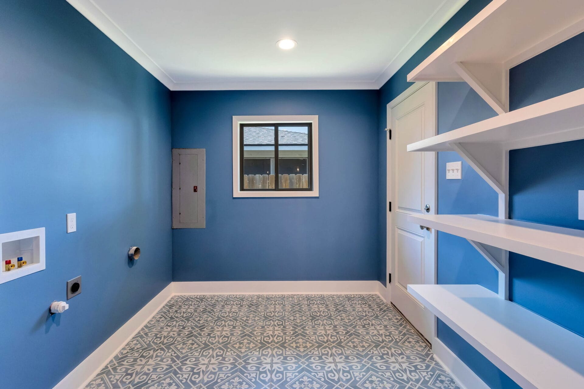 A laundry room with blue walls, patterned blue and white tile floor, open white shelves on the right, a window in the center, and plumbing and electrical hookups on the left wall.