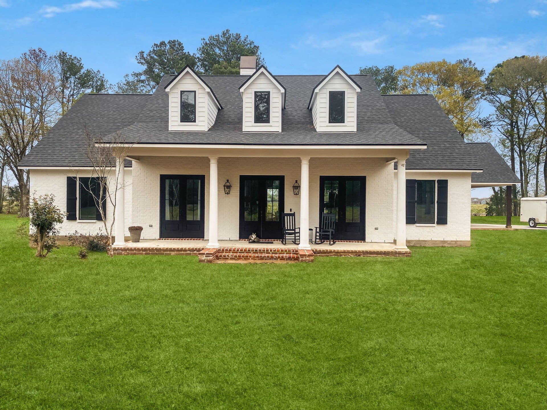 A white two-story house with black shutters and three dormer windows, featuring a covered front porch with two rocking chairs, sits on a large green lawn with trees in the background.