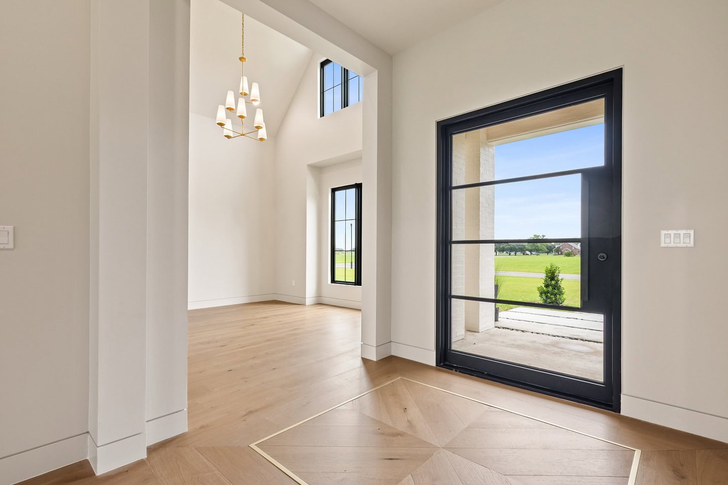 A modern entryway with light wood flooring, white walls, large black-framed windows, a geometric chandelier, and double glass doors opening to a covered patio with a view of grass and trees outside.