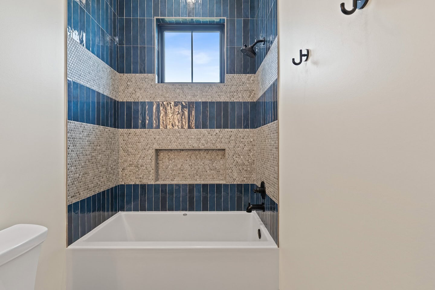 A white bathtub is set against a tiled wall with blue vertical tiles and beige mosaic horizontal stripes. A window and a built-in shelf are above the tub. Black fixtures and wall hooks are visible.