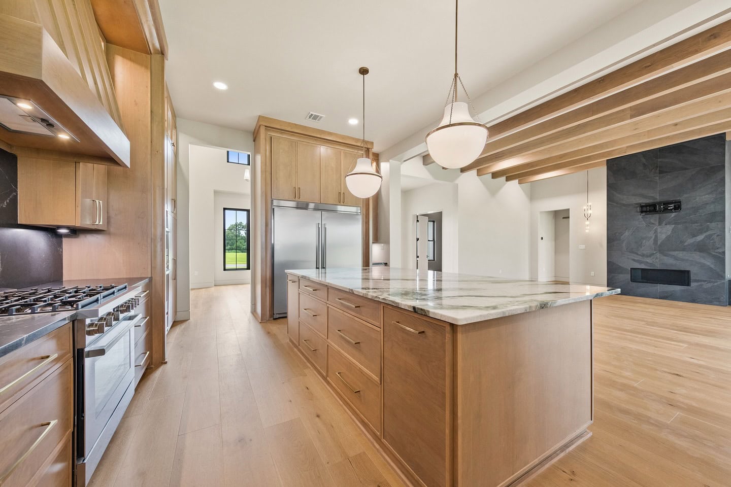 Modern kitchen with light wood cabinets, marble island countertop, stainless steel appliances, pendant lights, and open layout leading to a spacious living area with wood floors and beamed ceilings.