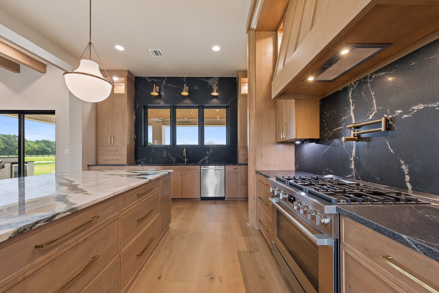Modern kitchen with wood cabinets, marble island, stainless steel appliances, and black marble backsplash. Large window above sink, pendant light over island, and view of outdoors through sliding glass door.