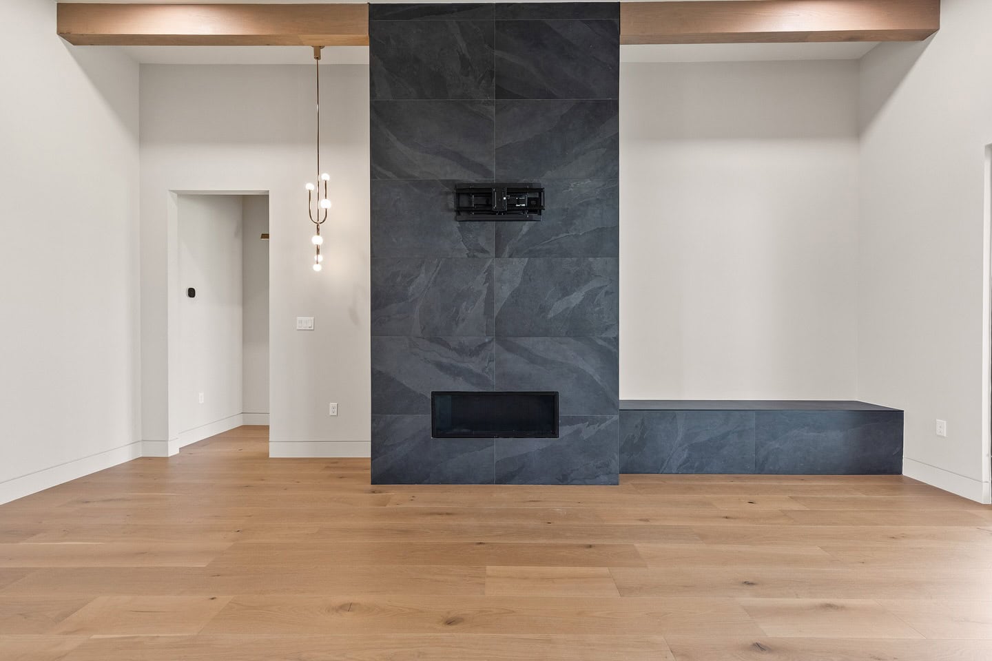 A modern living room with light wood flooring and a central dark slate accent wall featuring a built-in fireplace and bench, pendant light, and white walls with exposed wood ceiling beam.