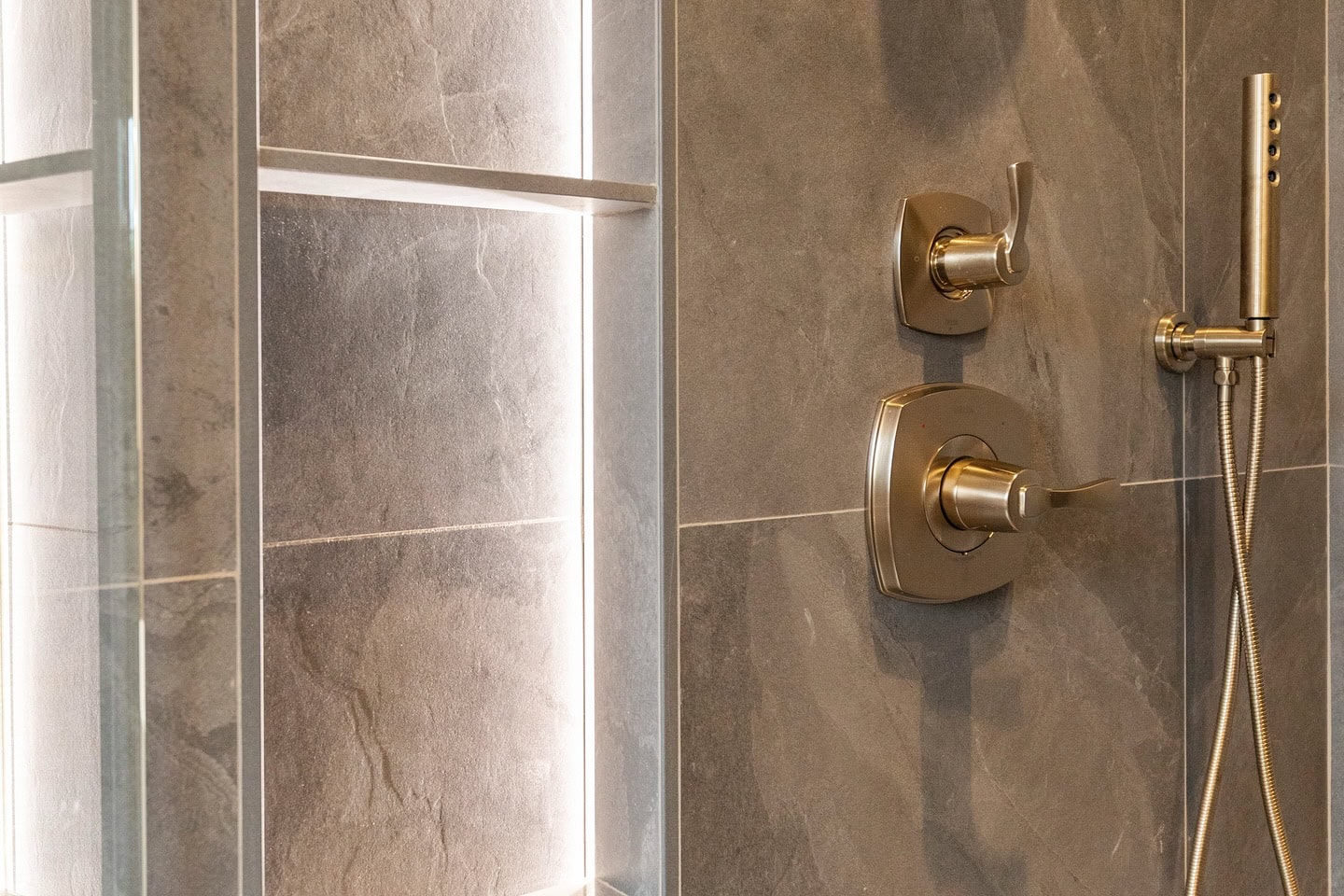 Close-up of a modern shower with gray stone tiles, chrome faucet handles, and a handheld showerhead mounted on the wall. Vertical lighting highlights the texture of the tiles.