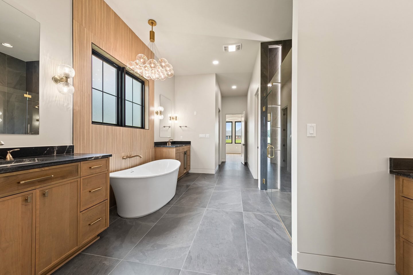 Modern bathroom with large gray floor tiles, a freestanding white bathtub, wood cabinets with black countertops, wall-mounted lights, and a glass shower enclosure. A hallway leads to another room with windows.