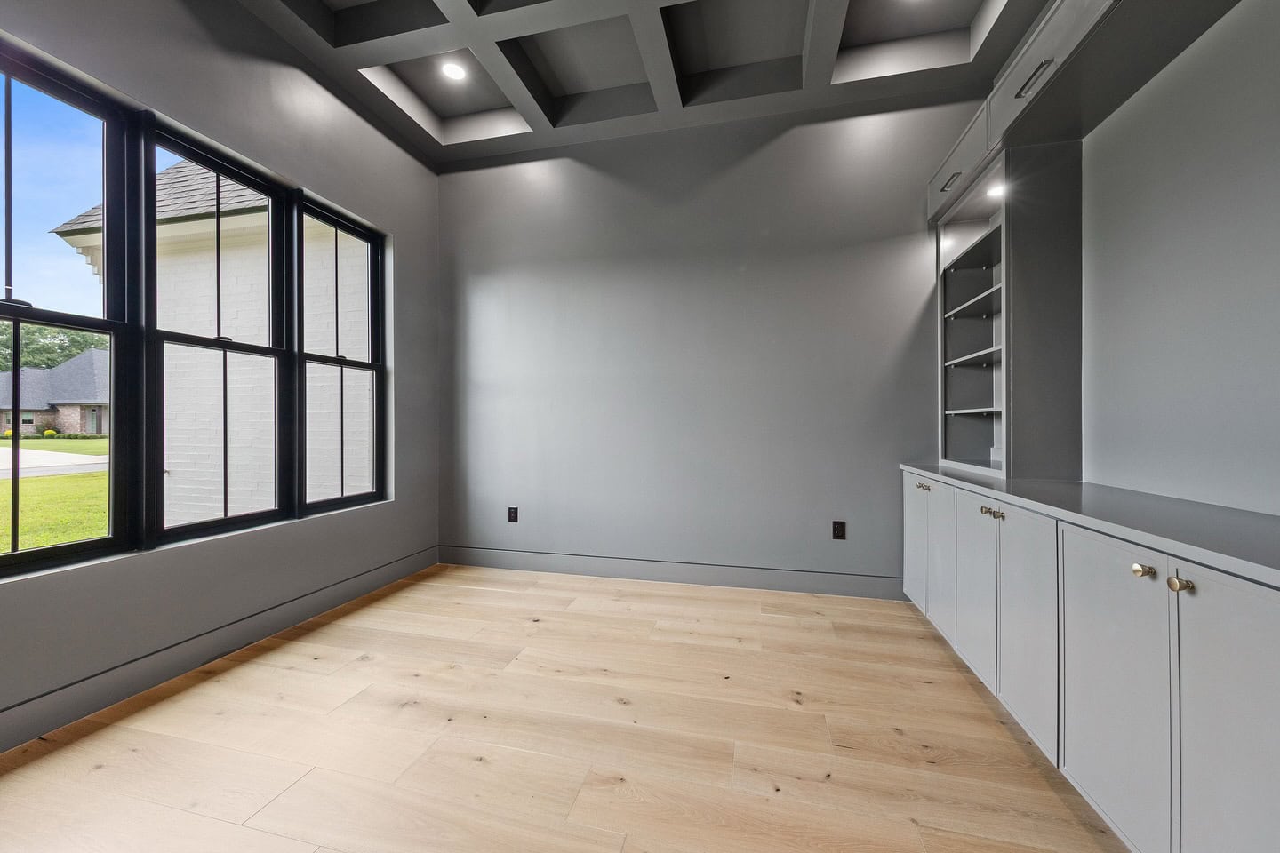 A modern, empty room with light wood flooring, built-in cabinets and shelves on one wall, large black-framed windows, and a coffered ceiling with recessed lights. The room is painted in shades of gray.