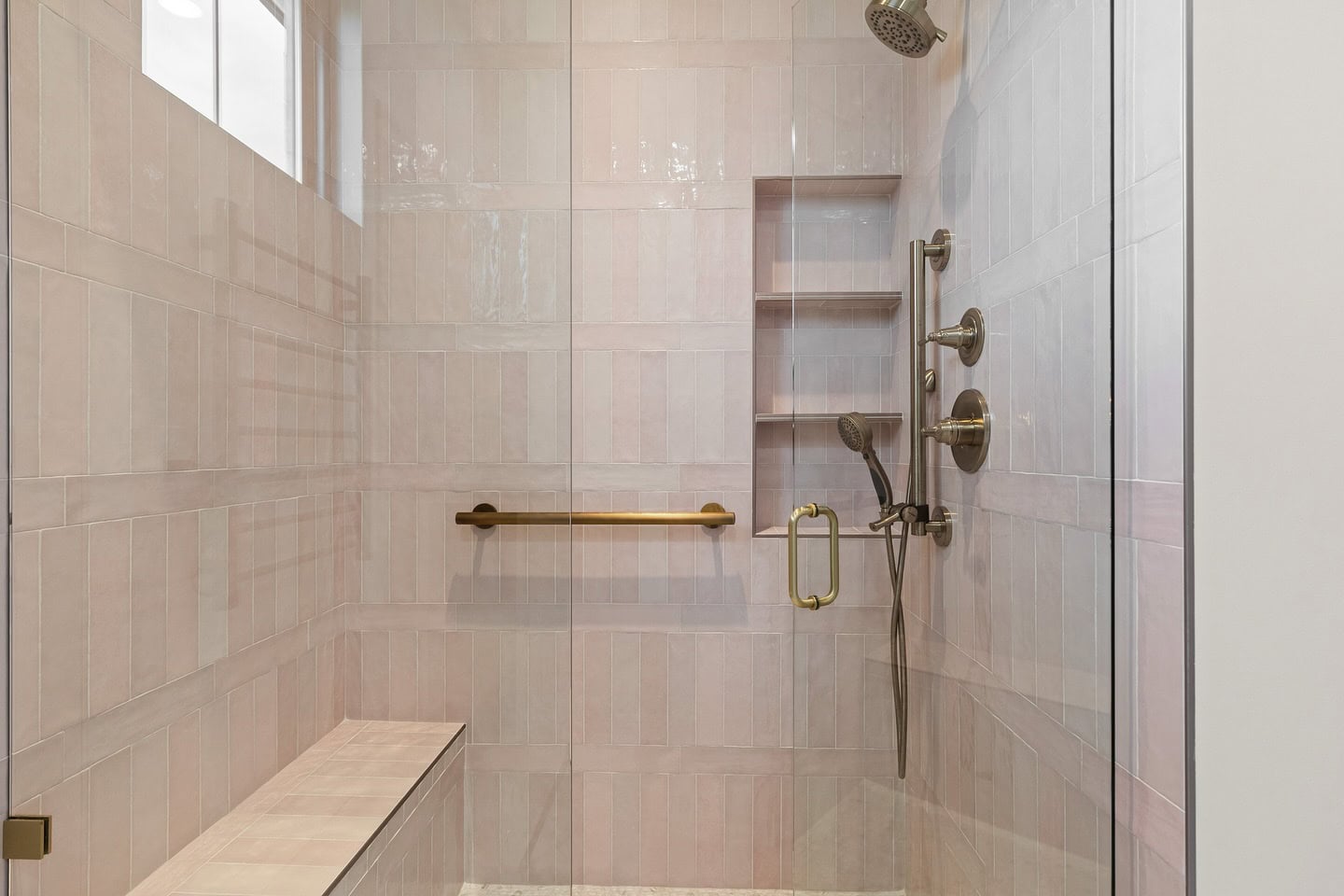 A modern walk-in shower with light pink rectangular tiles, a built-in bench, gold-colored grab bar and fixtures, a handheld showerhead, rainfall showerhead, and three recessed shelves.