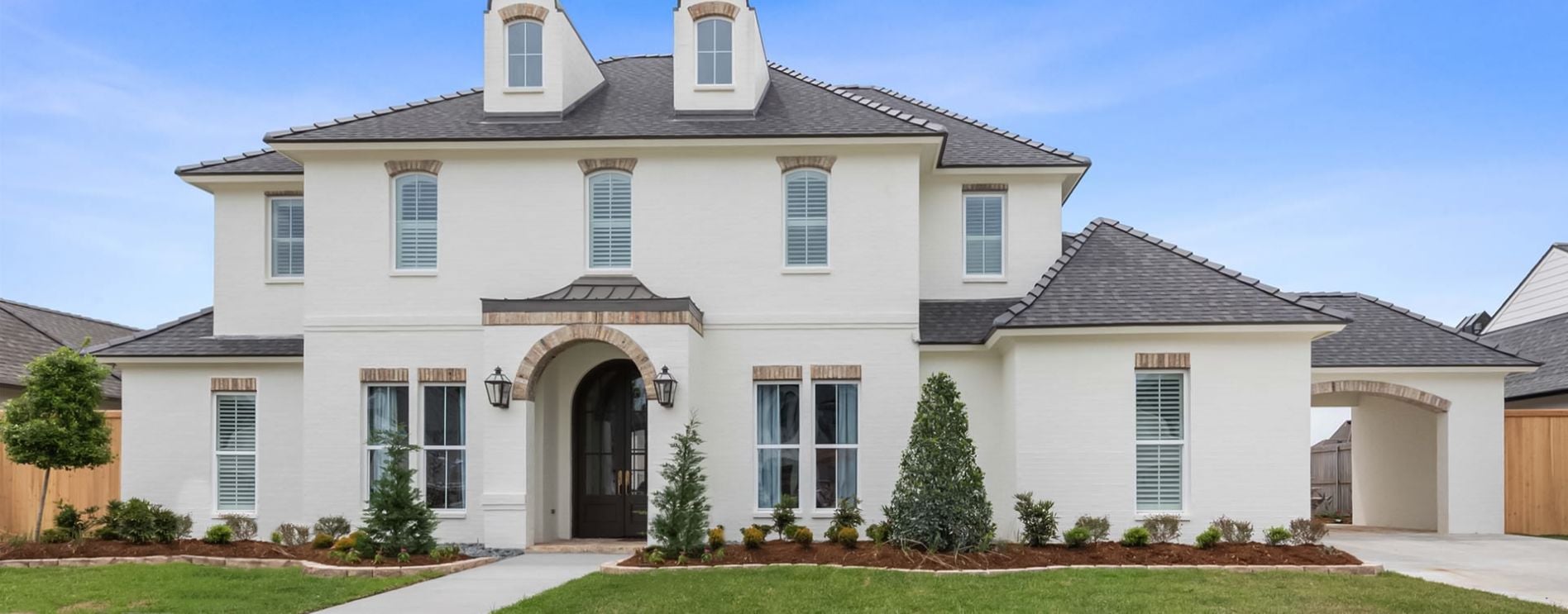 Two-story white brick house with gray roof, arched entryway, double front doors, dormer windows, attached carport, and landscaped front yard with small trees and bushes under a blue sky.