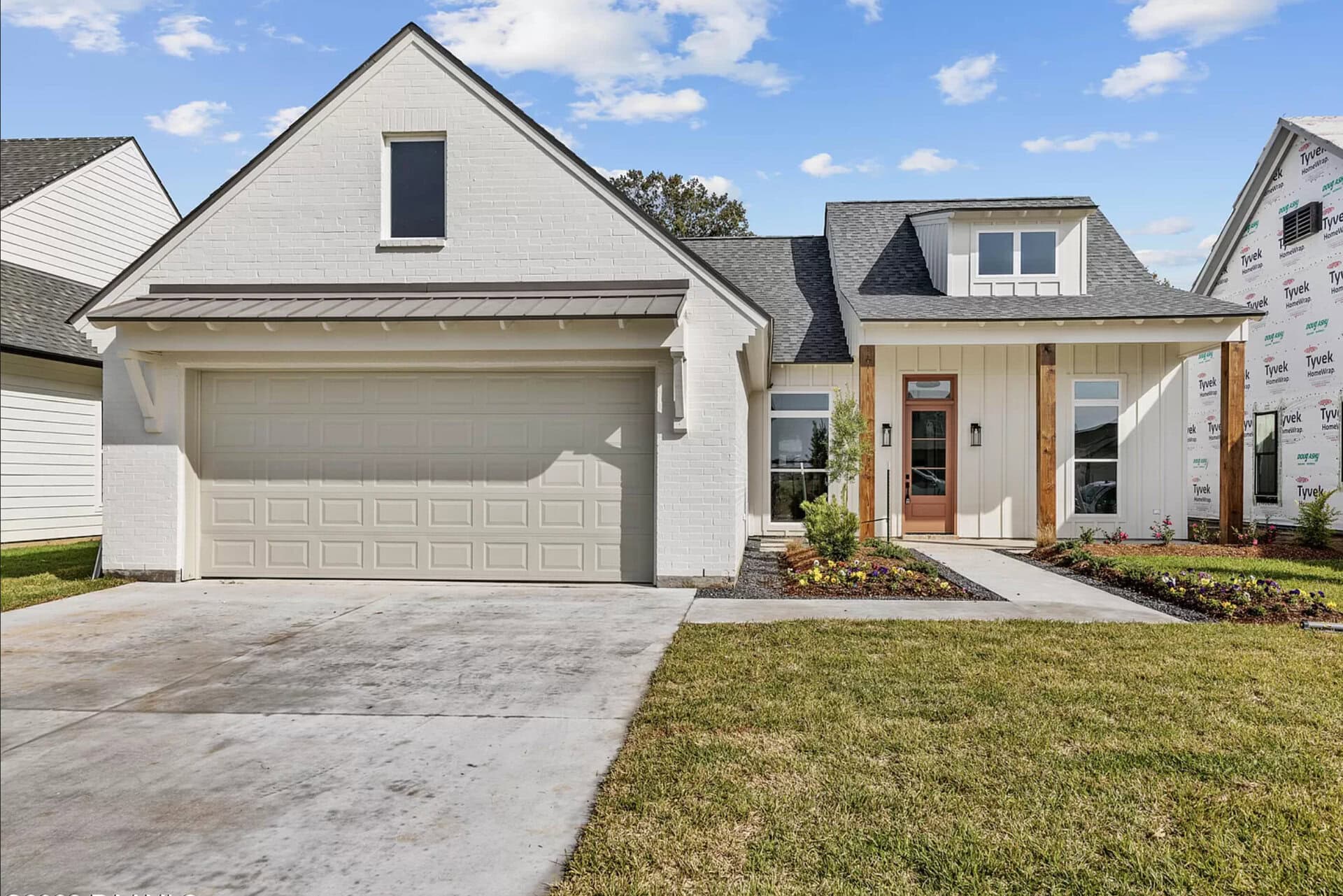 A modern single-story house with white brick exterior, attached two-car garage, small front porch with wooden columns, manicured lawn, and a concrete driveway under a partly cloudy sky.